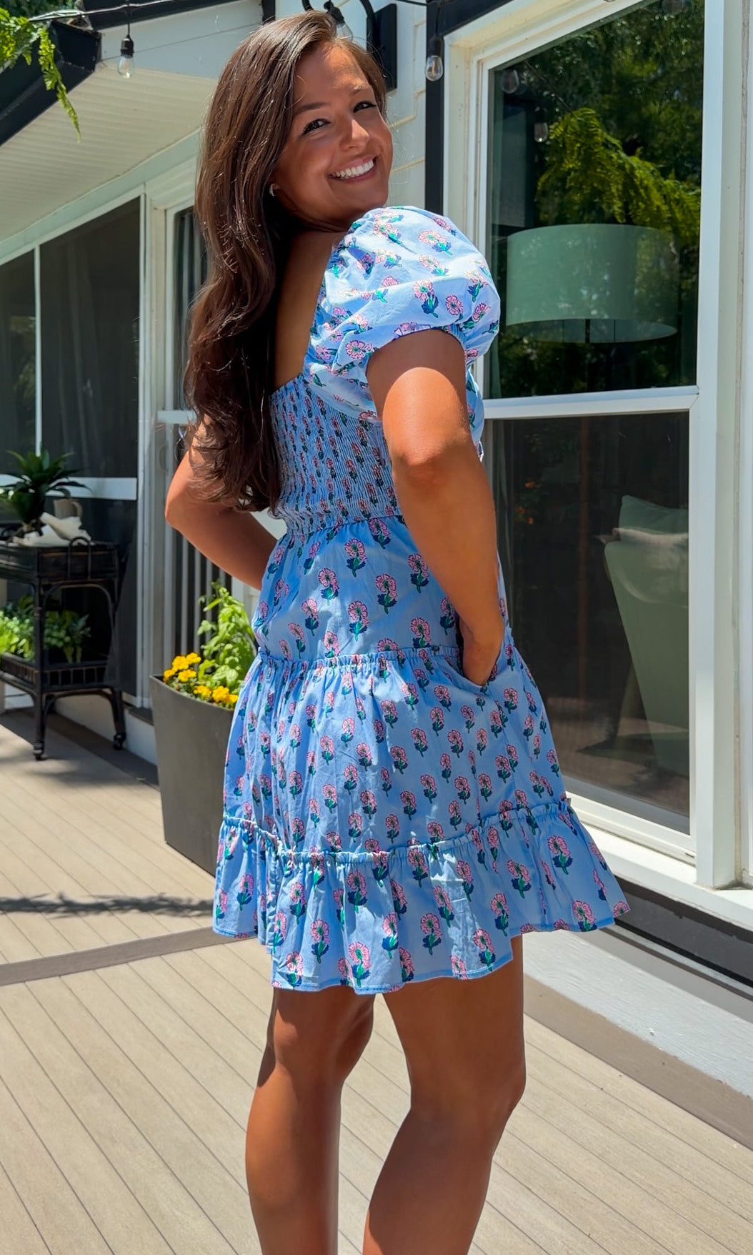 Woman in a blue dress standing on a deck with plants and a house in the background