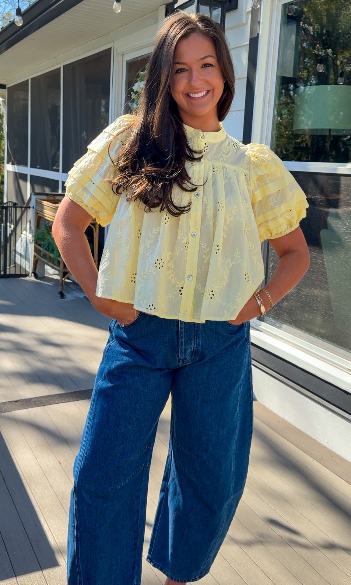 Woman wearing a yellow blouse and blue jeans standing on a wooden deck.