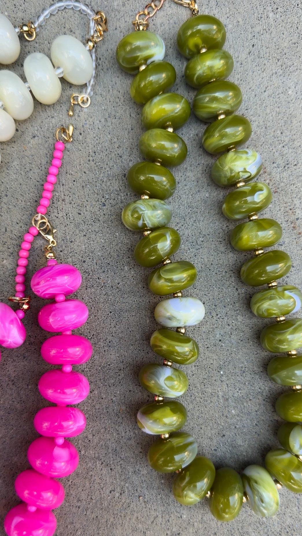 Three beaded necklaces laid flat on a gray textured surface: one with large olive green marbled beads, one with bright pink beads and white beads, and a partial third necklace with cream beads at the top left.