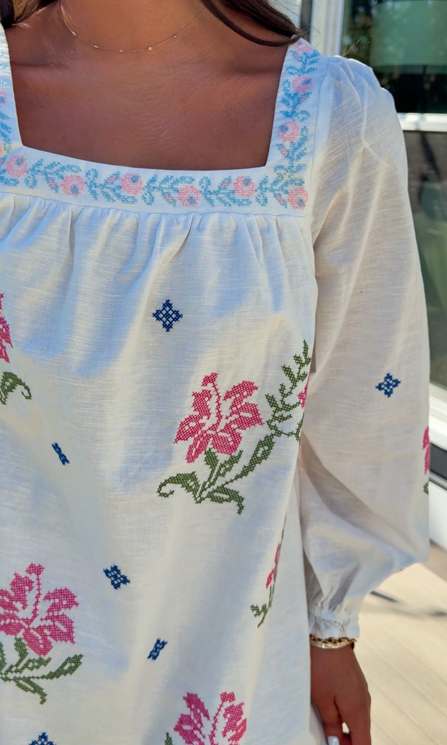 White embroidered mini dress with square neckline, long sleeves, and colorful floral embroidery, worn outdoors in natural light.