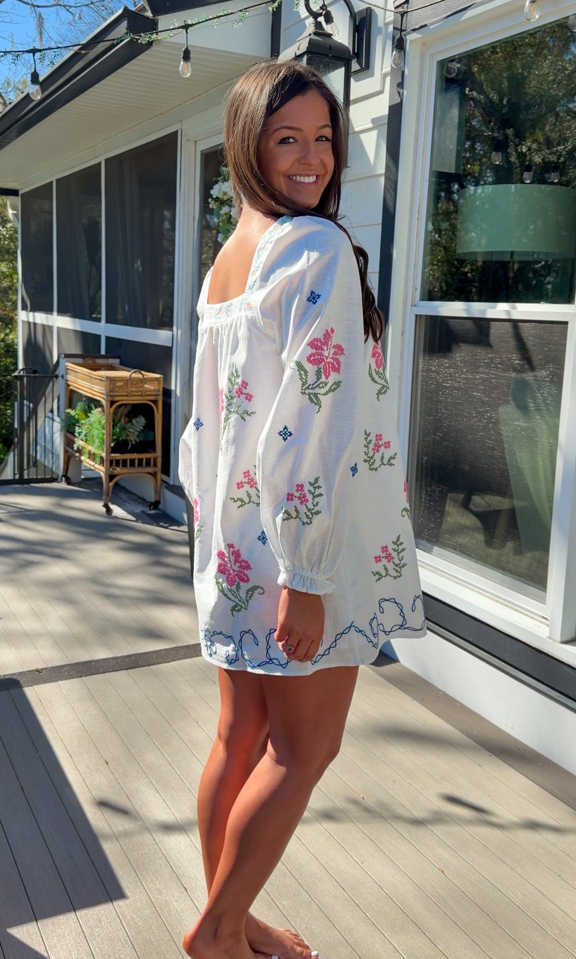 White embroidered mini dress with square neckline, long sleeves, and colorful floral embroidery, worn outdoors in natural light.