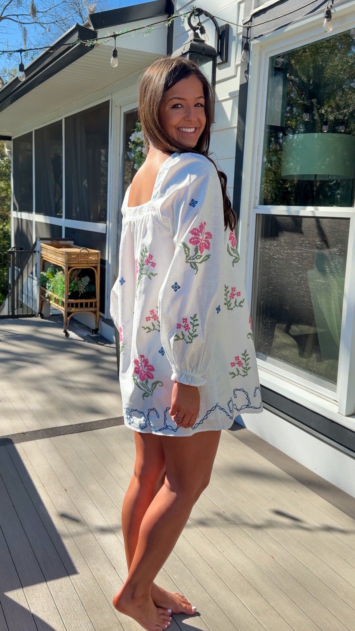 White embroidered mini dress with square neckline, long sleeves, and colorful floral embroidery, worn outdoors in natural light.