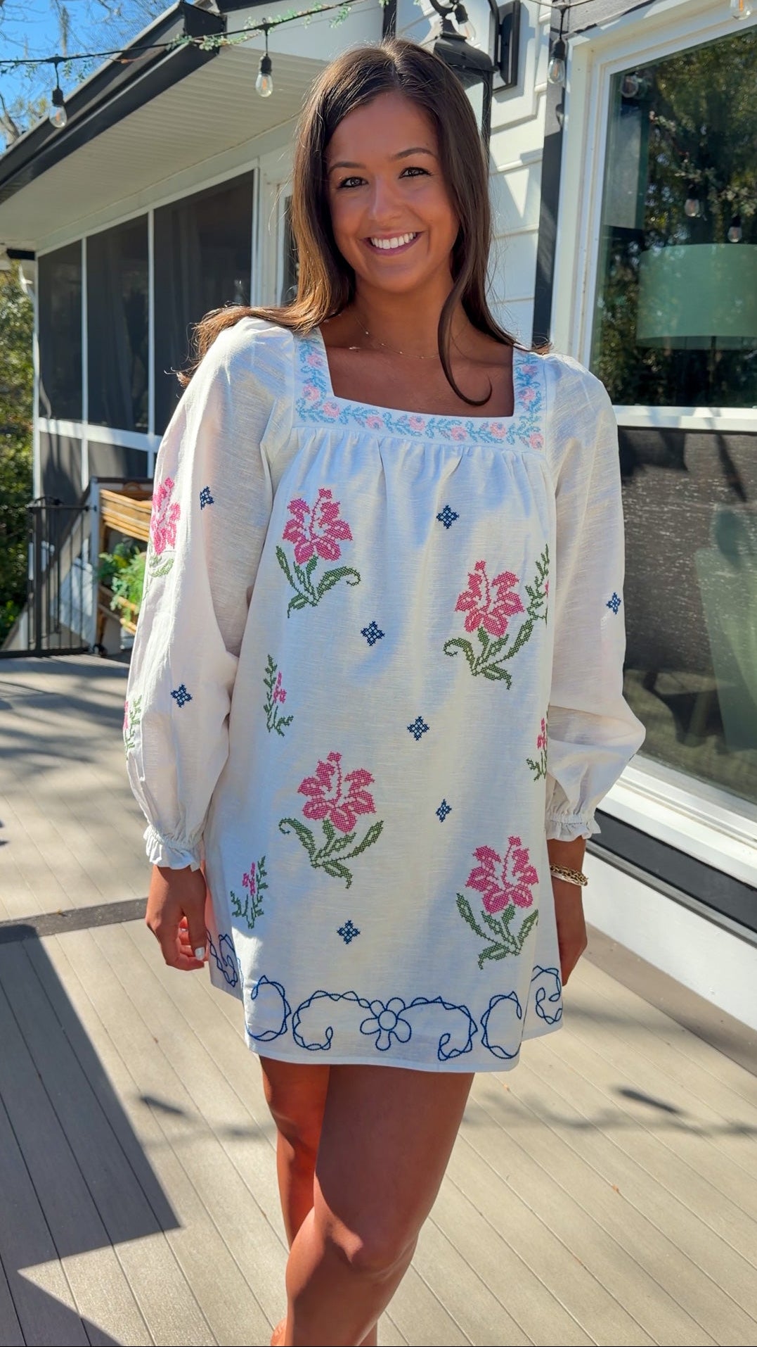 White embroidered mini dress with square neckline, long sleeves, and colorful floral embroidery, worn outdoors in natural light.