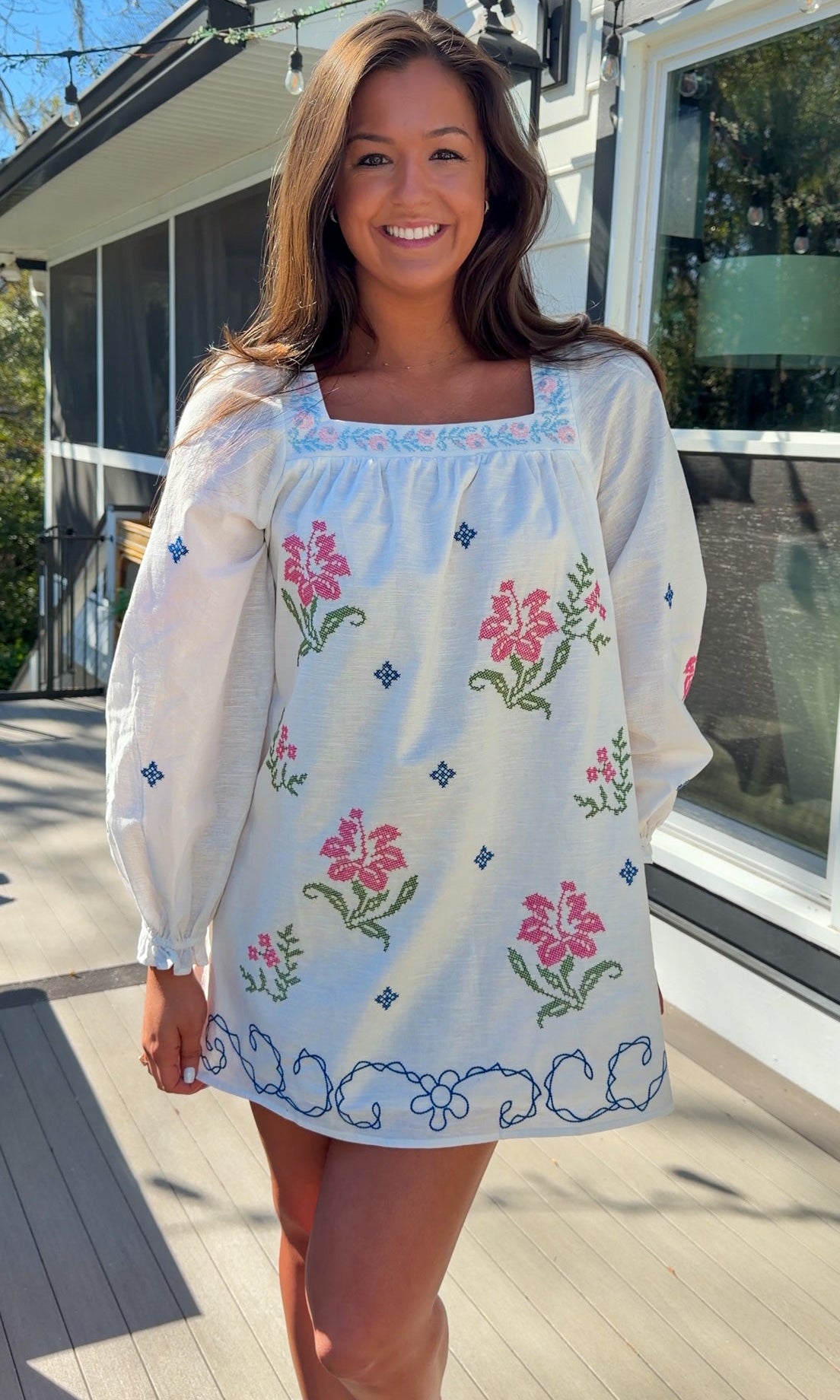 White embroidered mini dress with square neckline, long sleeves, and colorful floral embroidery, worn outdoors in natural light.