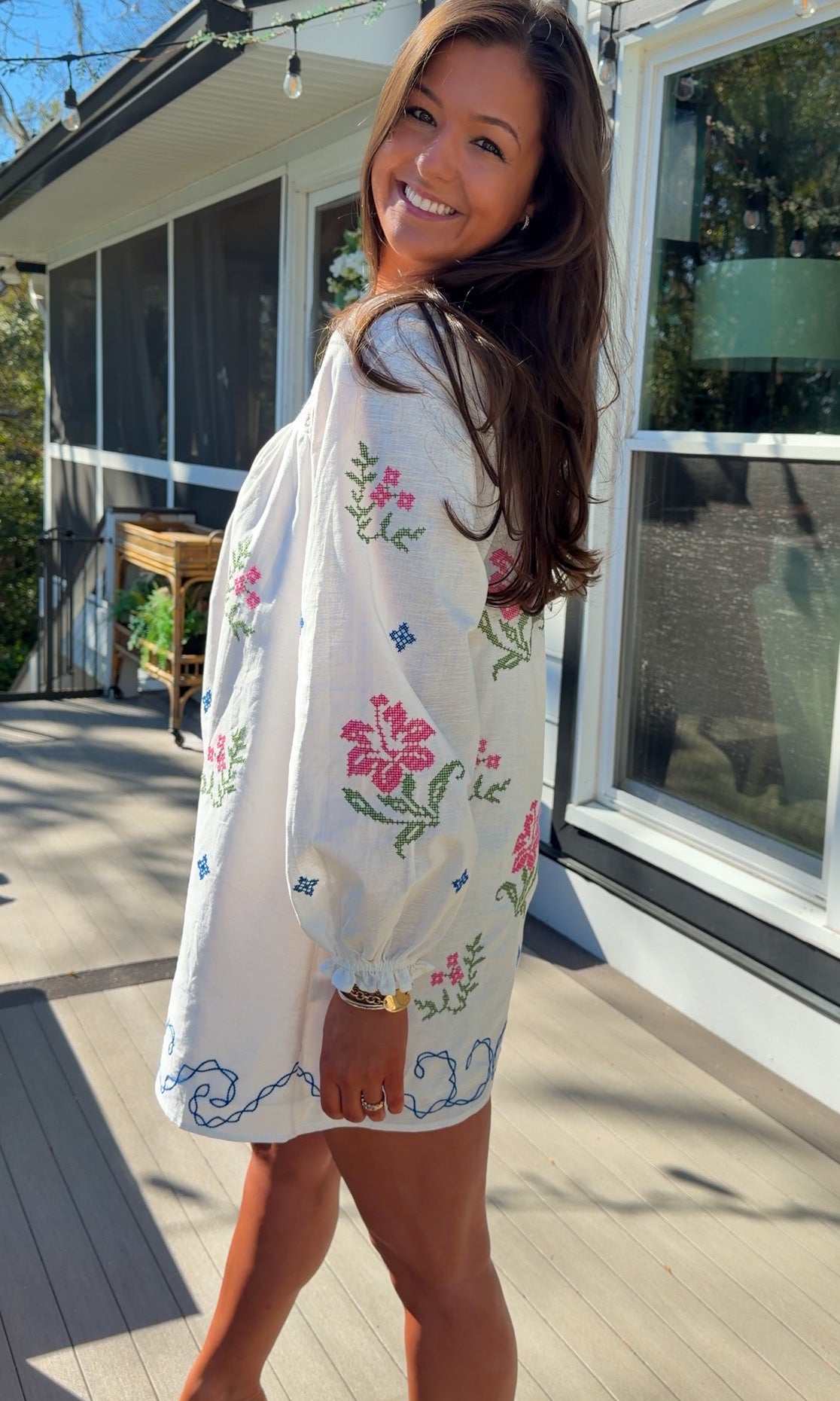 White embroidered mini dress with square neckline, long sleeves, and colorful floral embroidery, worn outdoors in natural light.