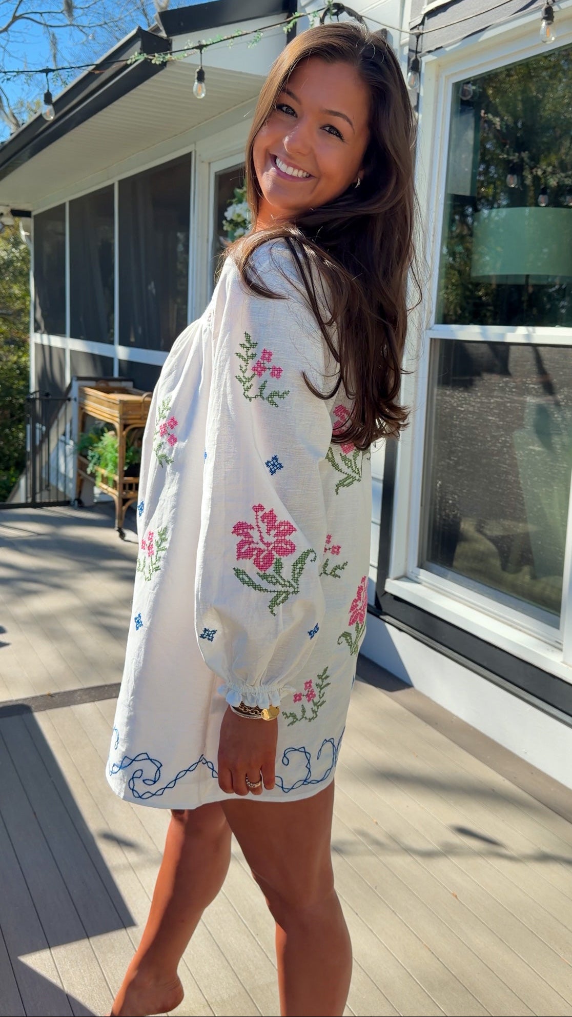 White embroidered mini dress with square neckline, long sleeves, and colorful floral embroidery, worn outdoors in natural light.