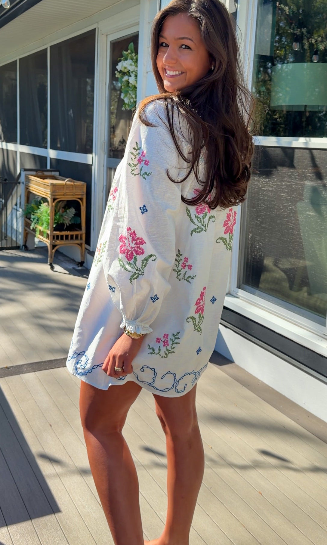 White embroidered mini dress with square neckline, long sleeves, and colorful floral embroidery, worn outdoors in natural light.