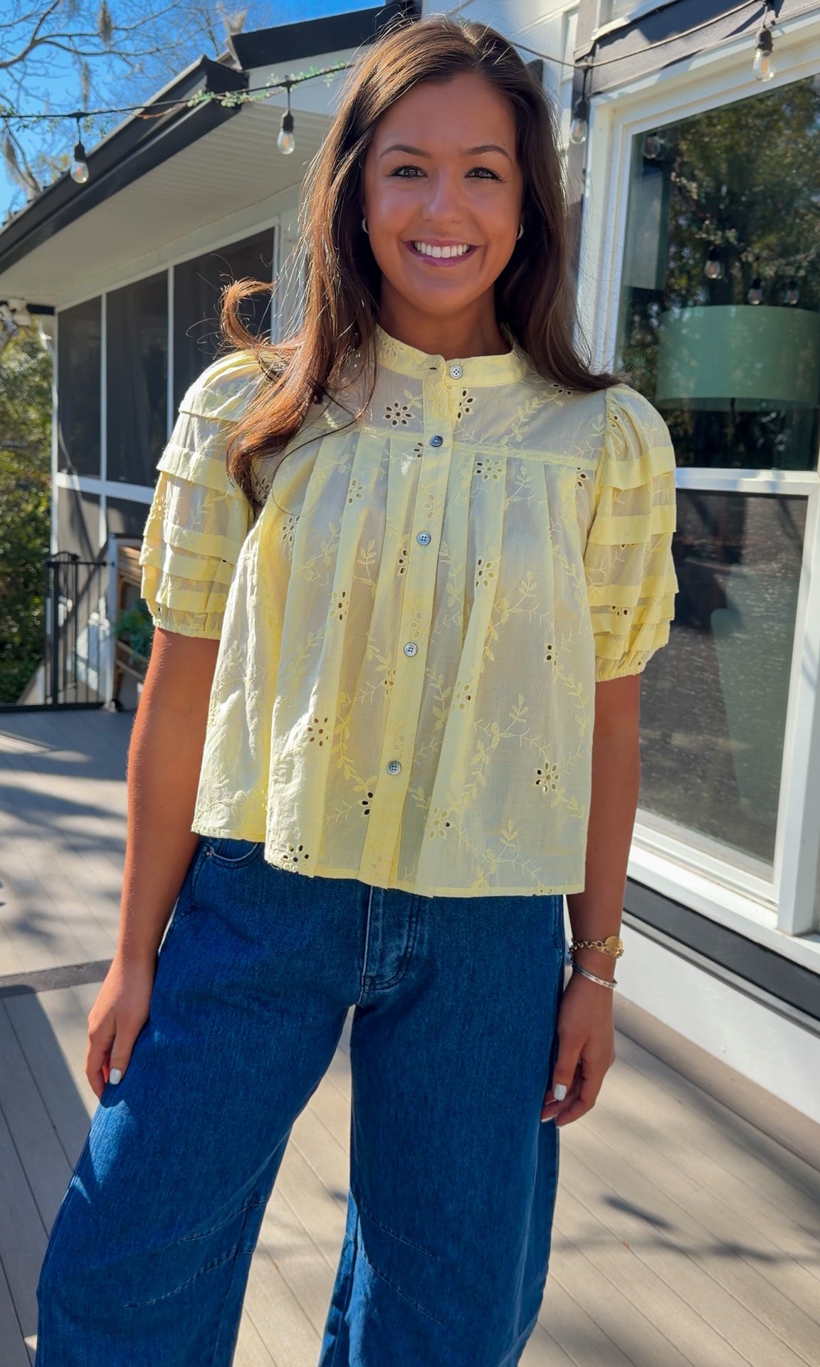 Woman wearing a yellow blouse with floral details and blue jeans standing outside a building.