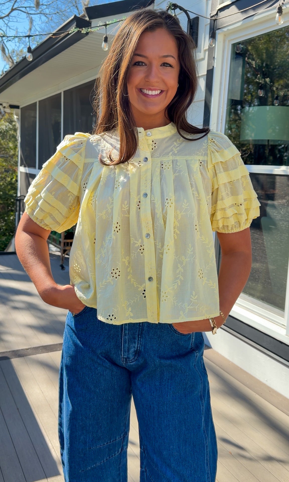 Woman wearing a yellow blouse with a pattern and blue jeans standing on a wooden deck.