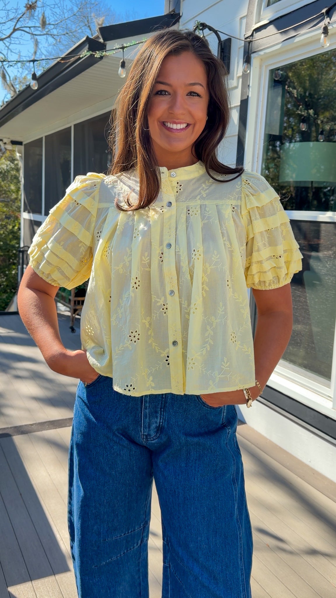Woman wearing a yellow blouse with a pattern and blue jeans standing on a wooden deck.