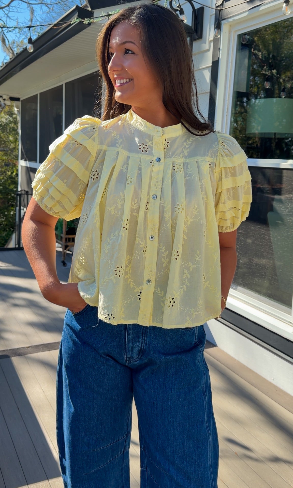 Woman wearing a yellow blouse with floral patterns and blue jeans standing outdoors.
