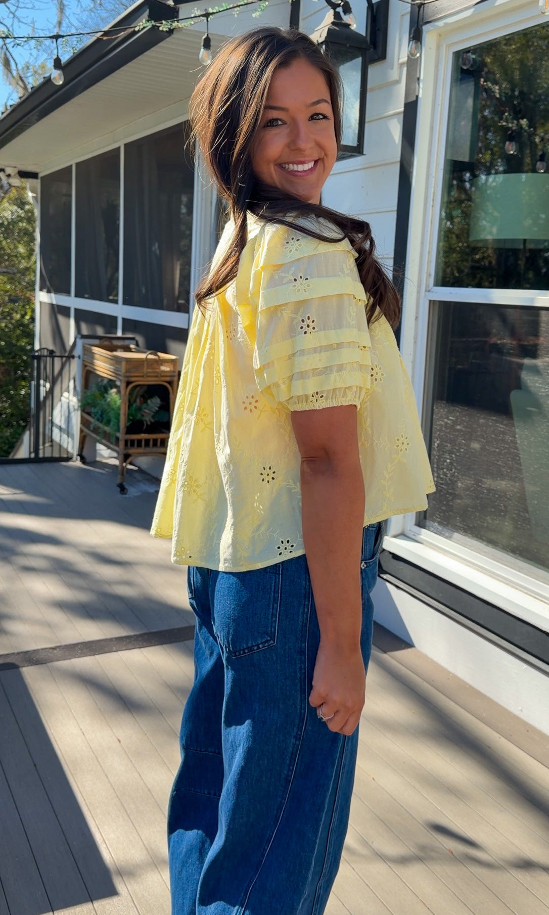 Woman wearing a yellow blouse and blue jeans standing on a wooden deck.