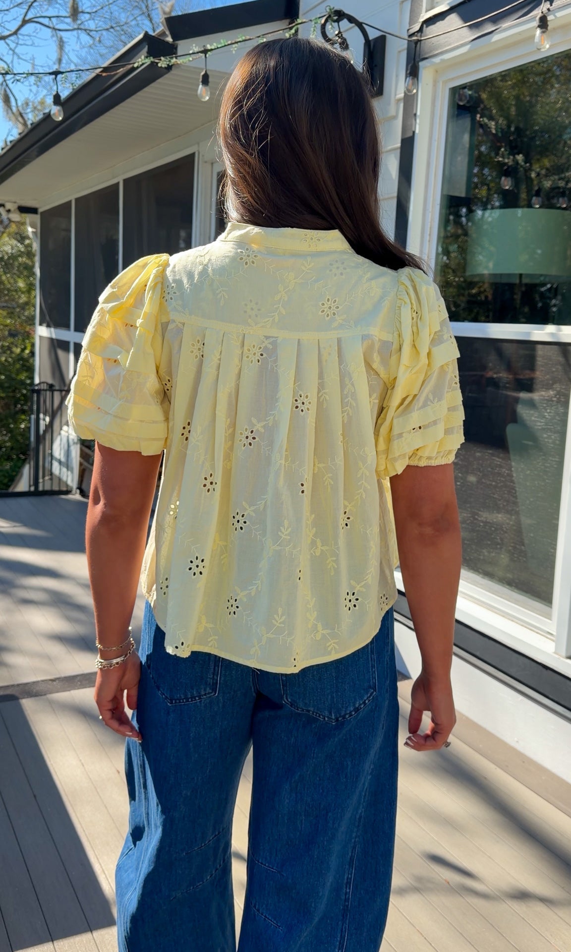 Person wearing a yellow blouse with floral patterns and blue jeans standing on a wooden deck.