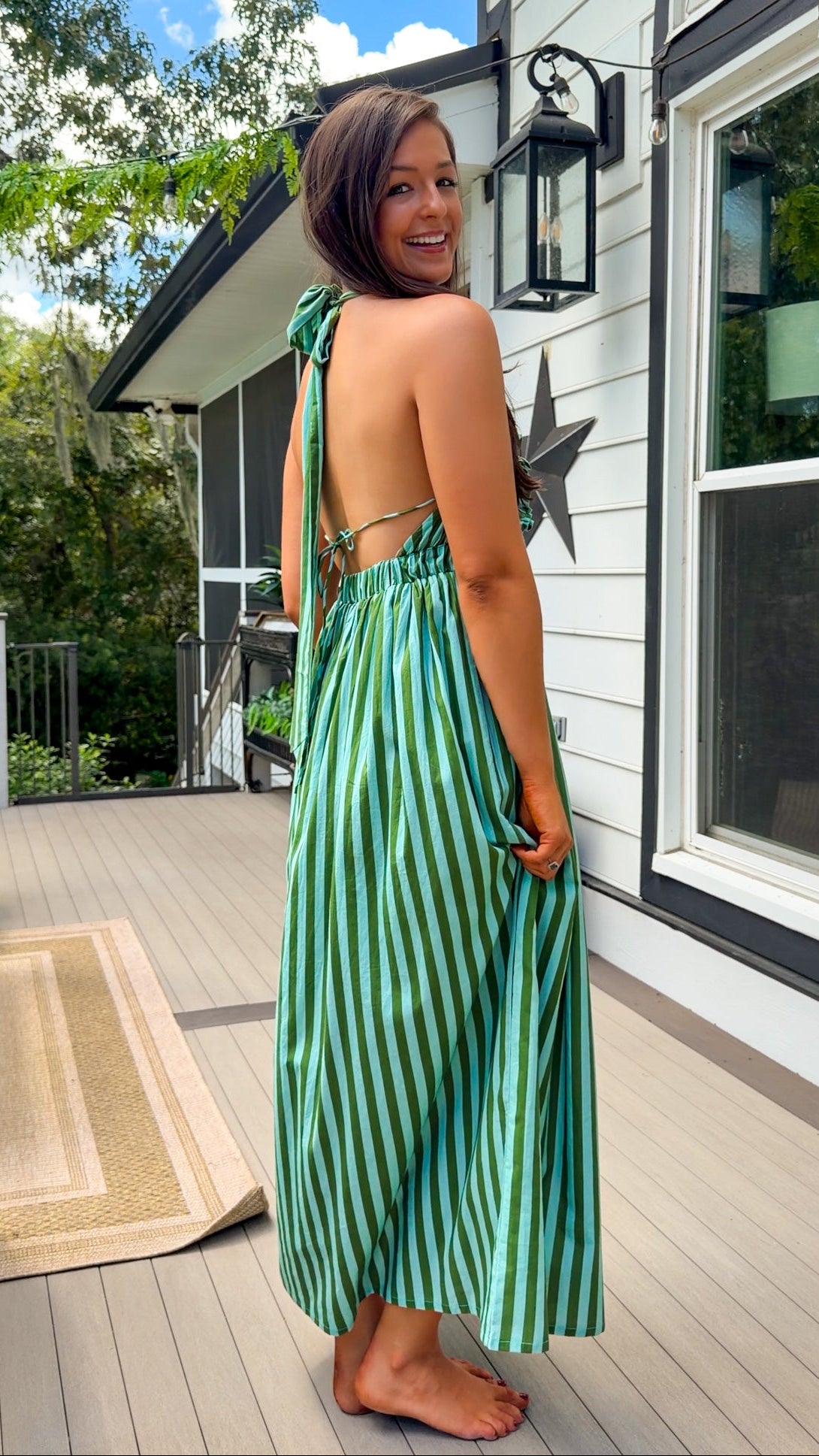 A woman wearing a green and teal striped halter maxi dress stands on a wooden deck in front of a white house, looking back over her shoulder.