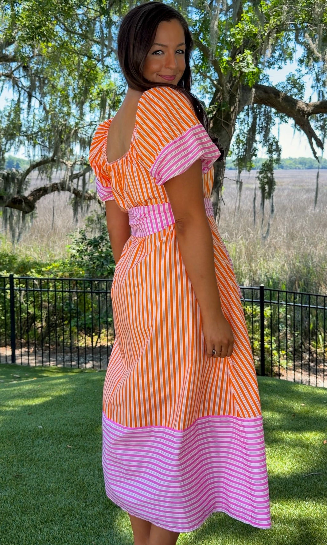 Woman wearing an orange and pink dress standing in a park with trees and grass.