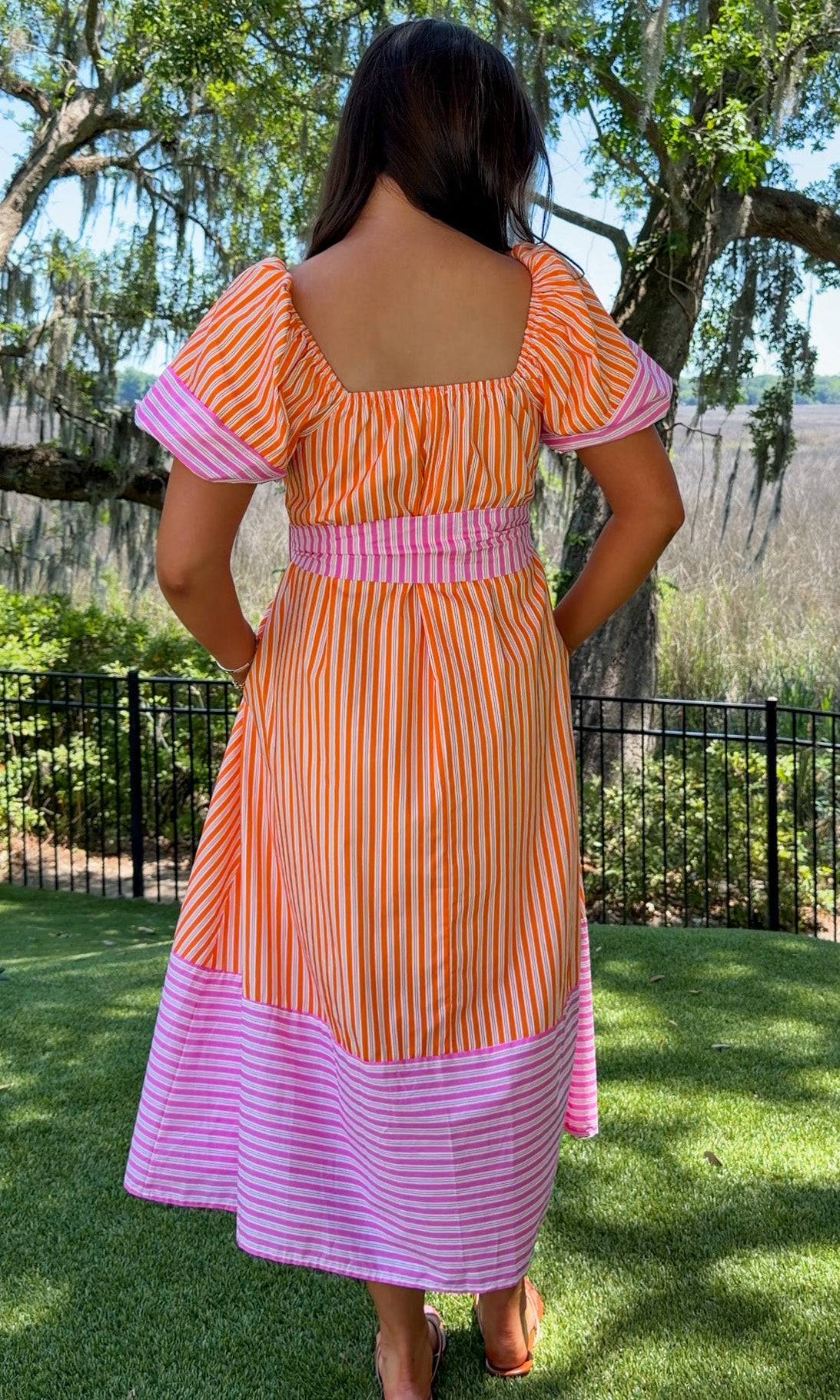 Woman wearing a colorful dress with a striped pattern, standing outdoors with trees and grass in the background.