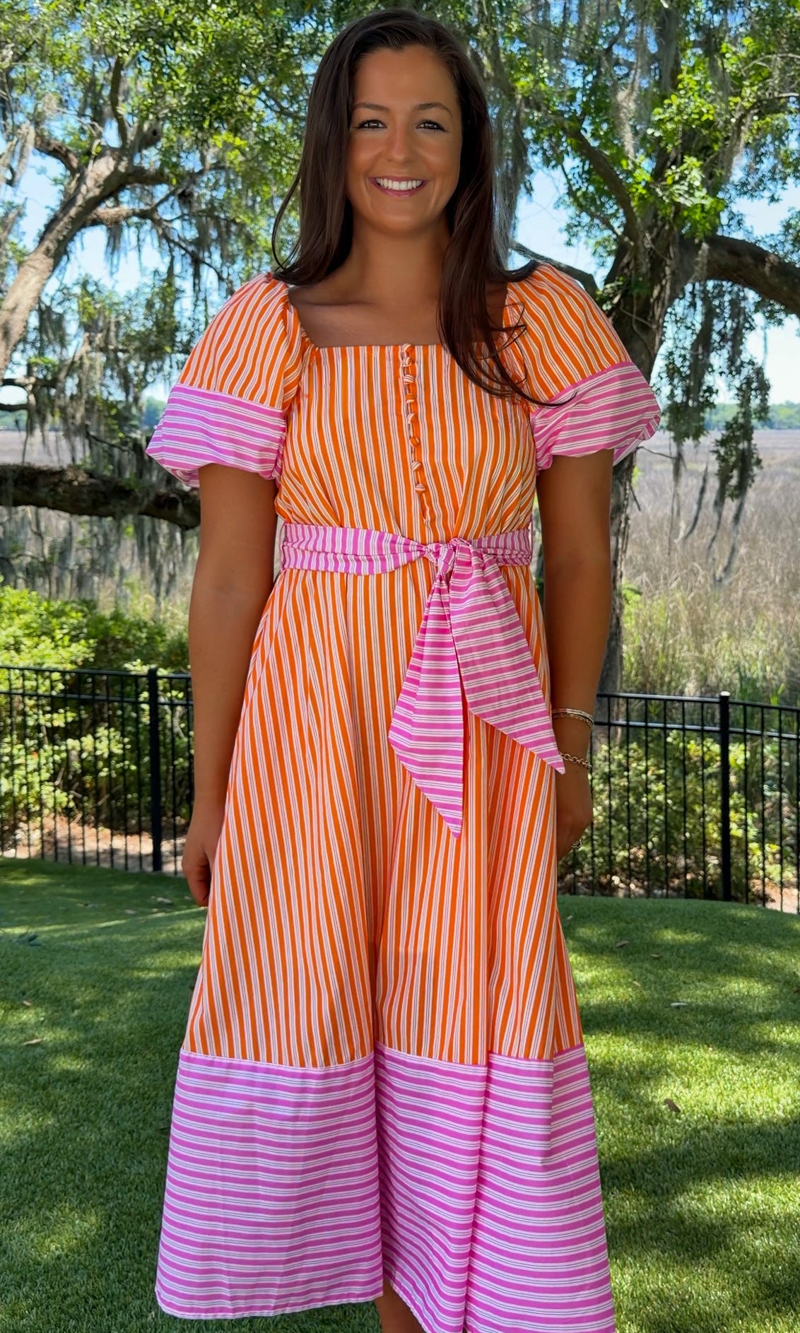 Woman wearing an orange and pink striped dress standing outdoors with trees and grass in the background