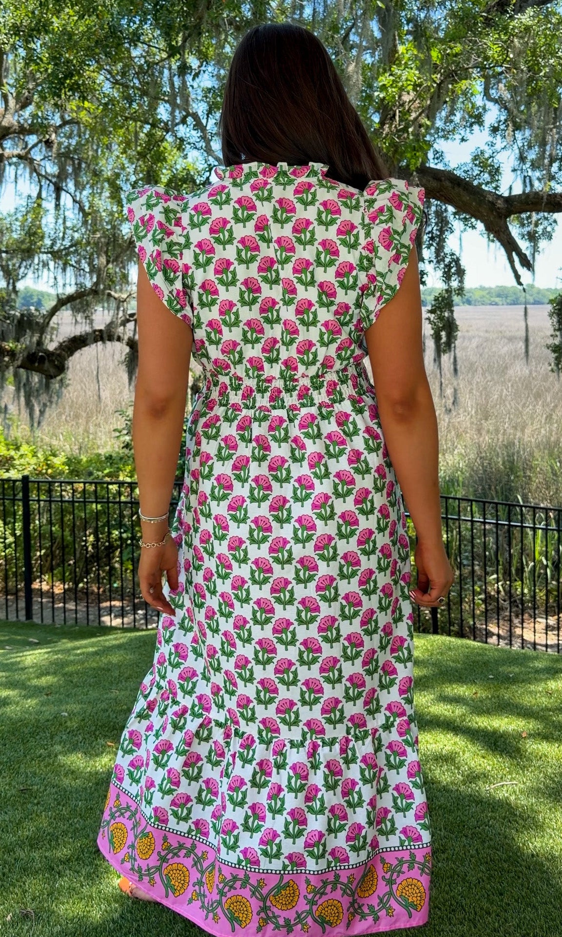 Woman wearing a floral dress standing in a garden with trees and greenery.