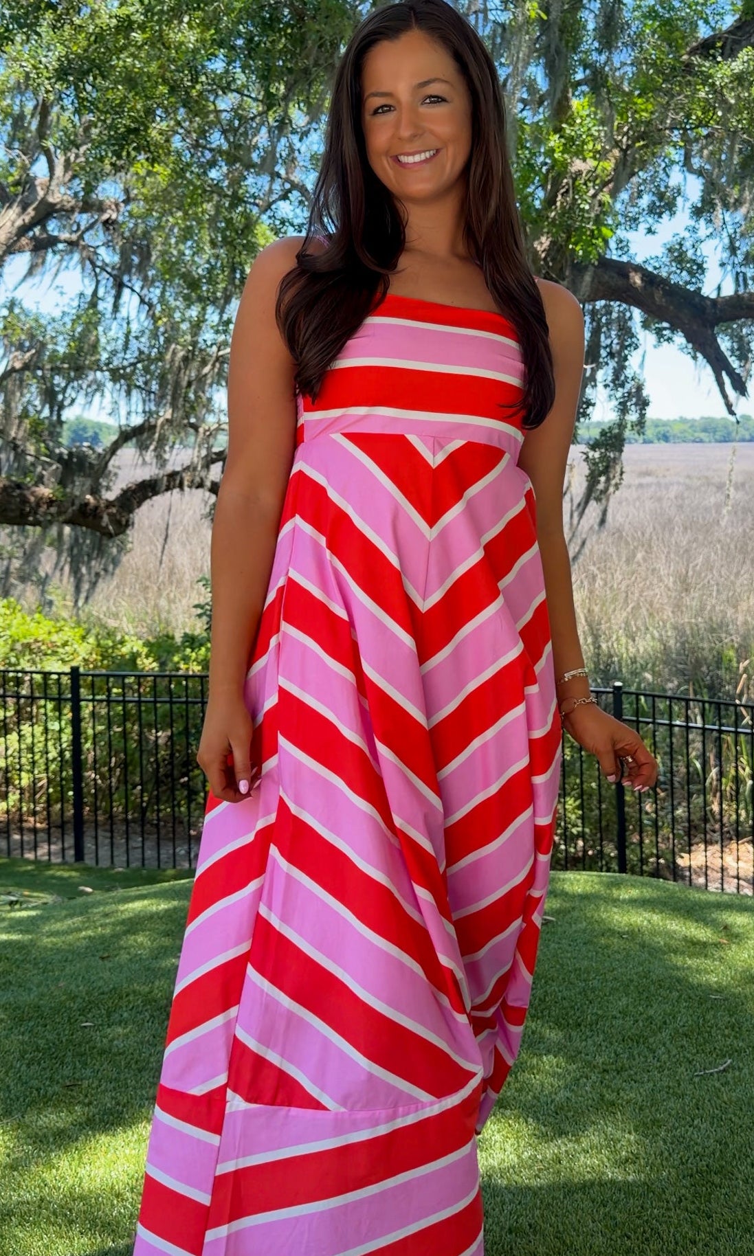 Woman wearing a red and pink striped dress standing outdoors with trees and grass in the background