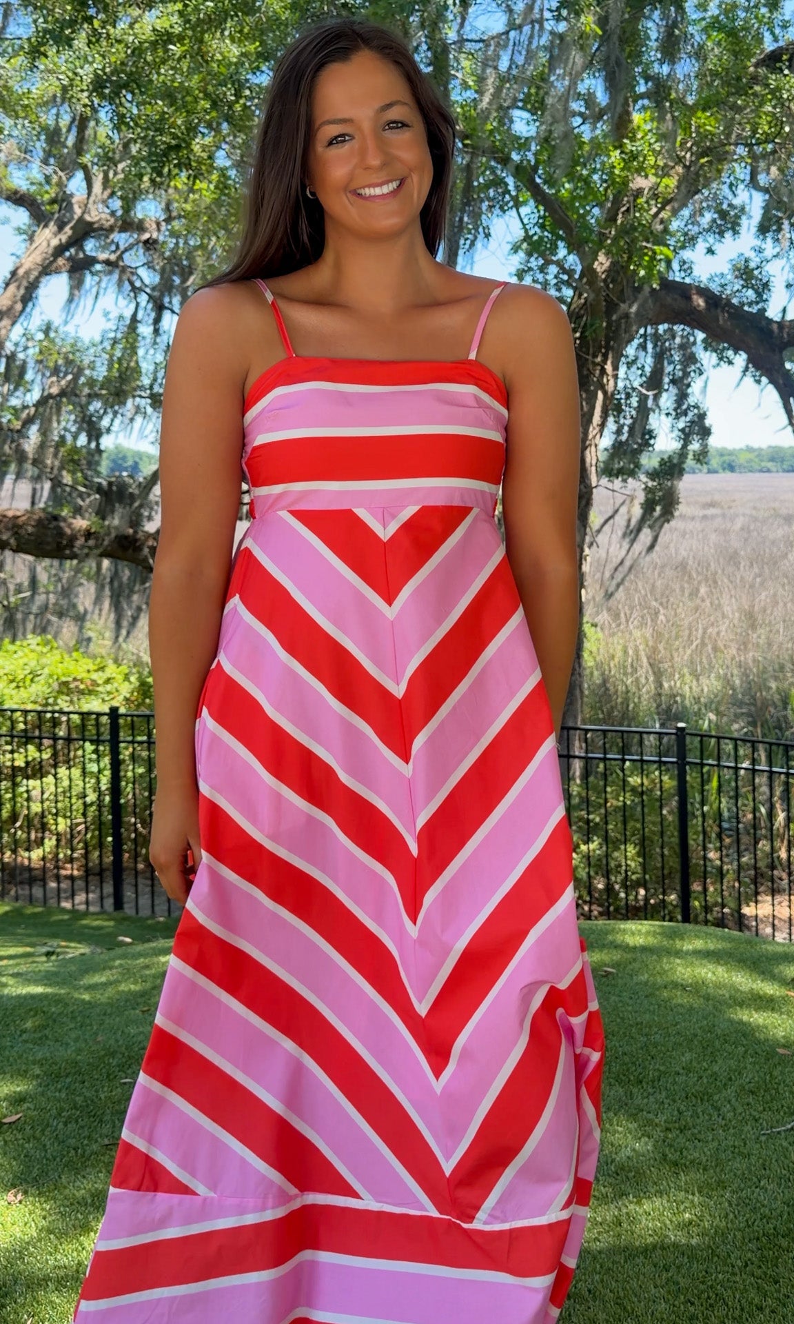 Woman wearing a red and pink striped dress standing outdoors with trees and grass in the background