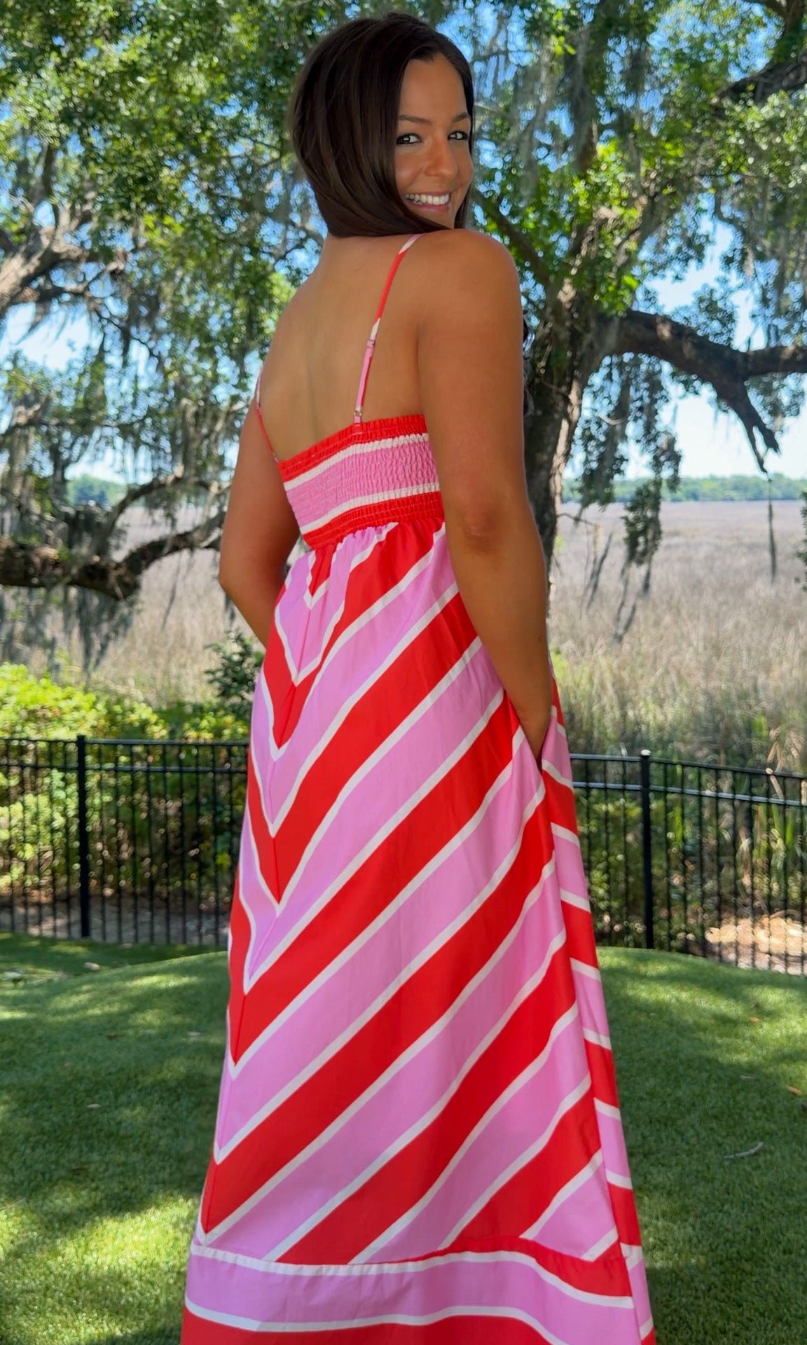 Woman wearing a red and pink striped dress standing outdoors with trees and grass in the background