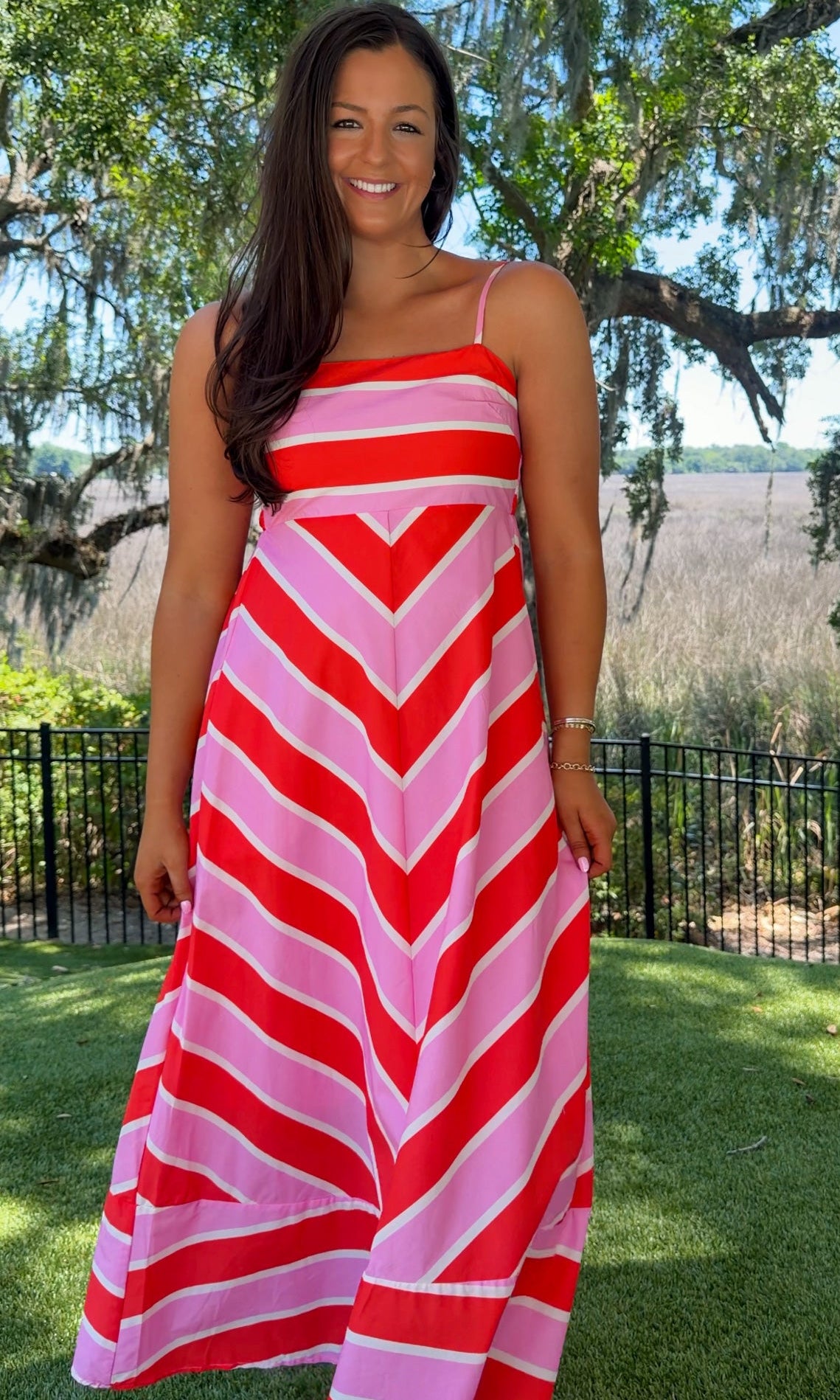 Woman wearing a red and pink striped dress standing outdoors with trees and grass in the background