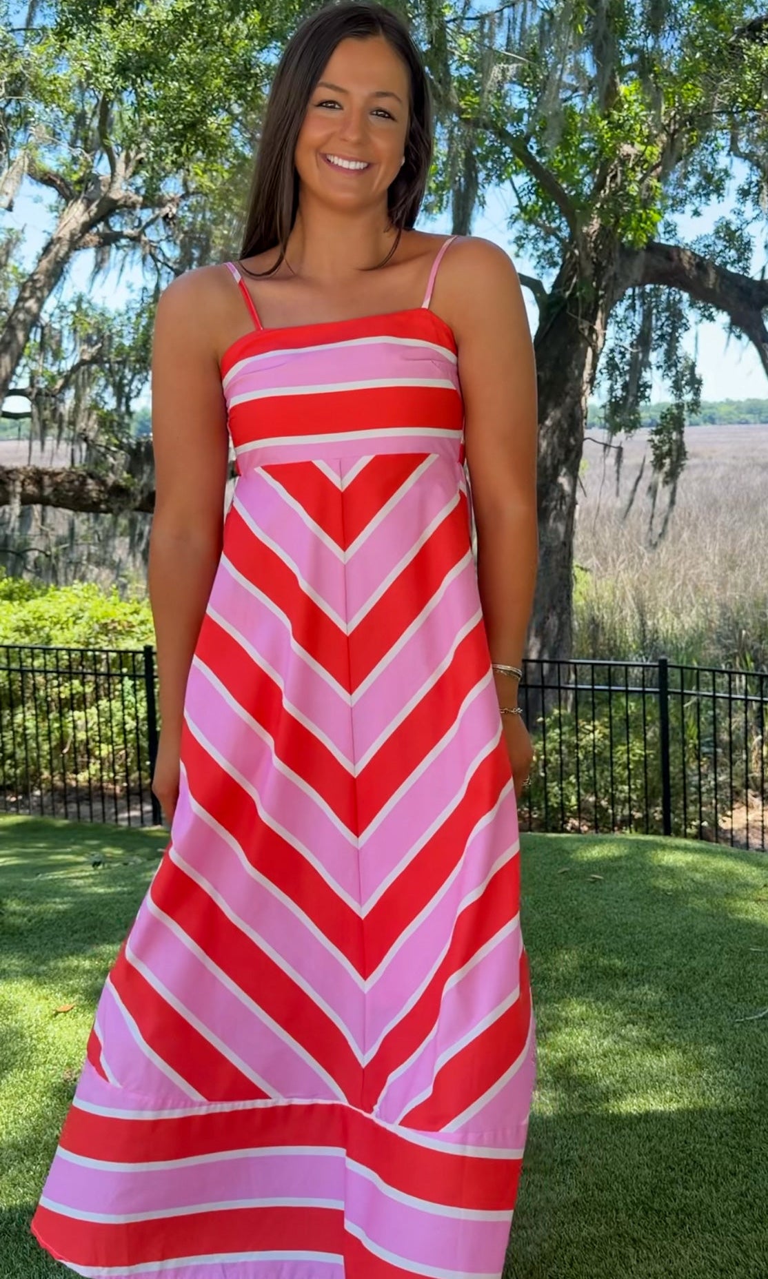 Woman wearing a red and pink striped dress standing outdoors with trees and grass in the background