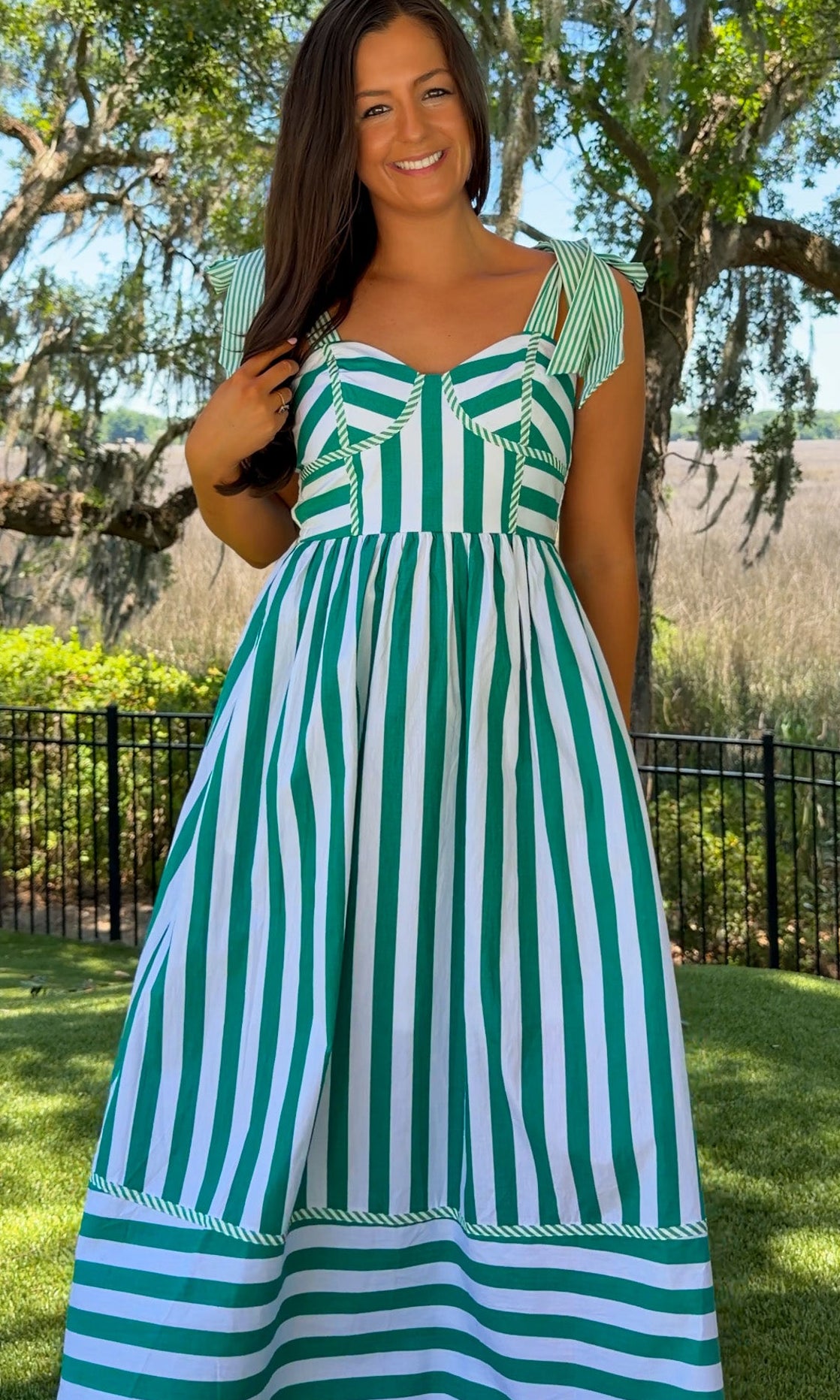 Woman wearing a green and white striped dress standing outdoors with trees and grass in the background