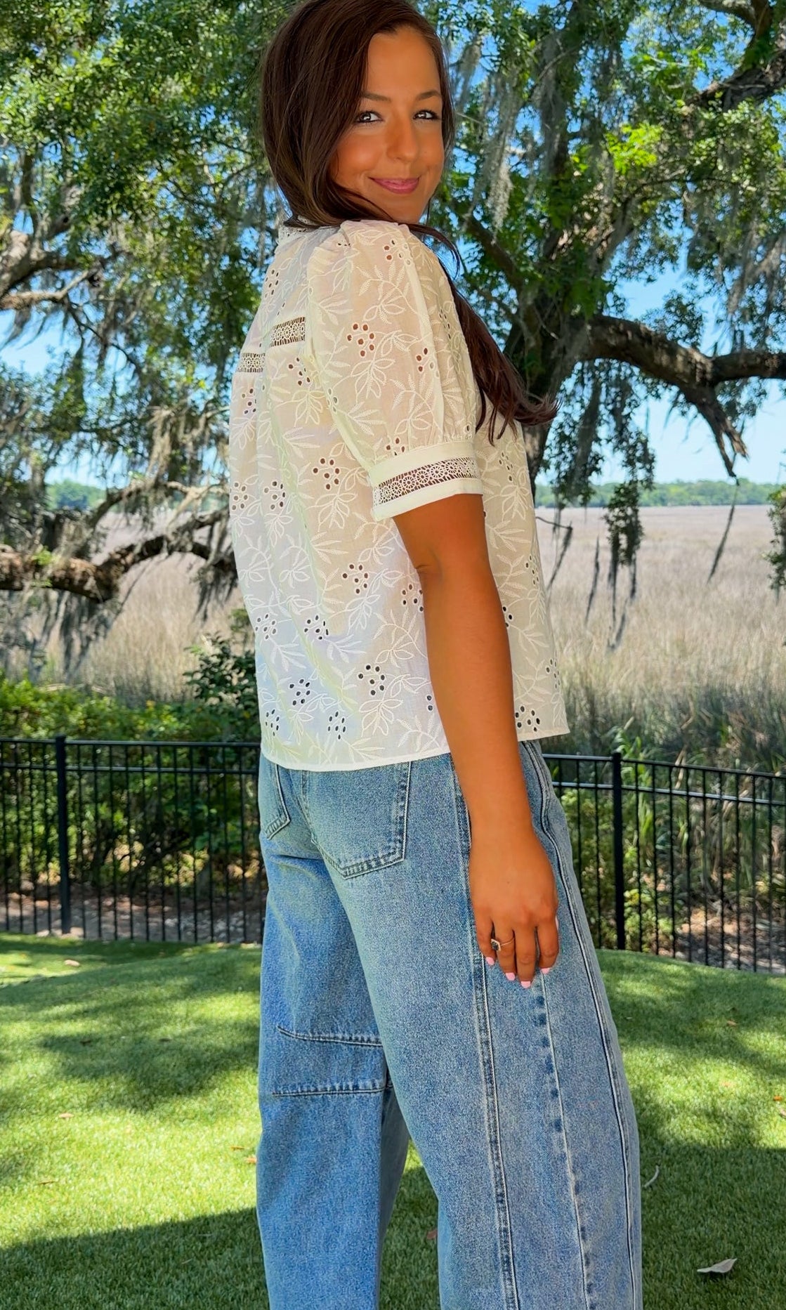 Woman wearing a white lace top and blue jeans standing outdoors with trees and grass in the background