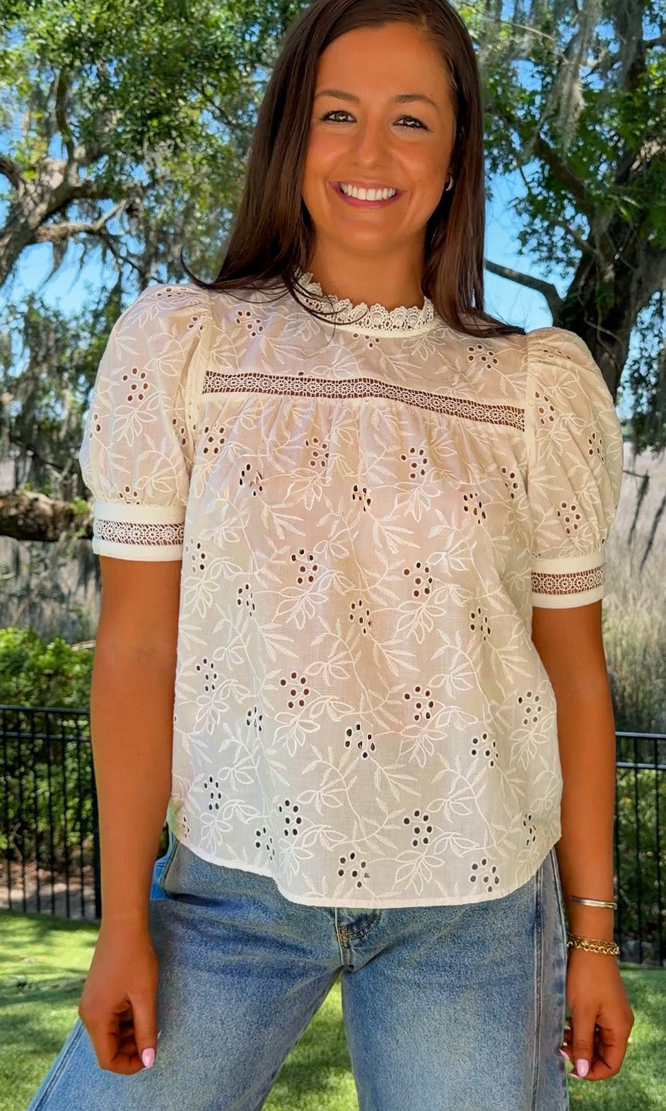Woman wearing a white lace top and blue jeans standing outdoors with trees and grass in the background
