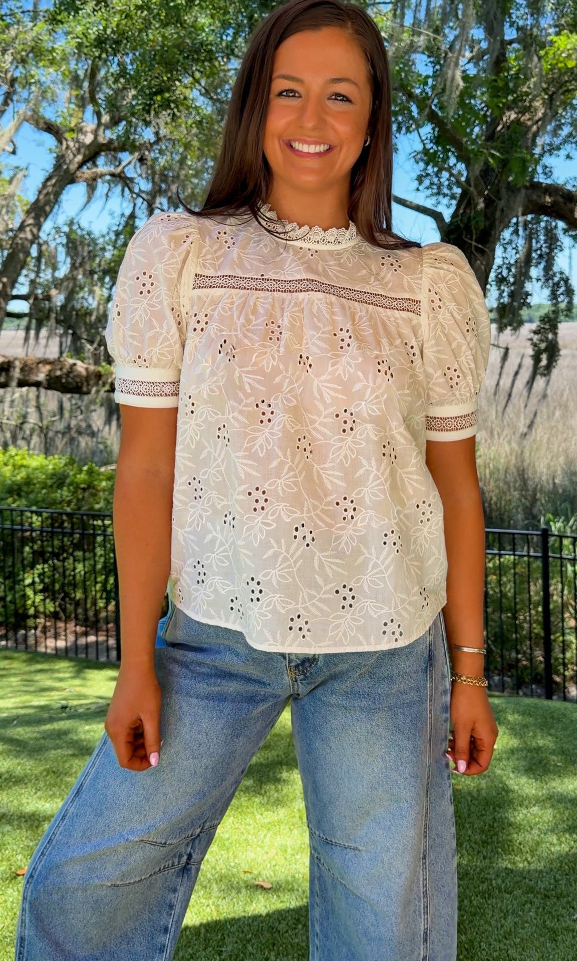Woman wearing a white lace top and blue jeans standing outdoors with trees and grass in the background