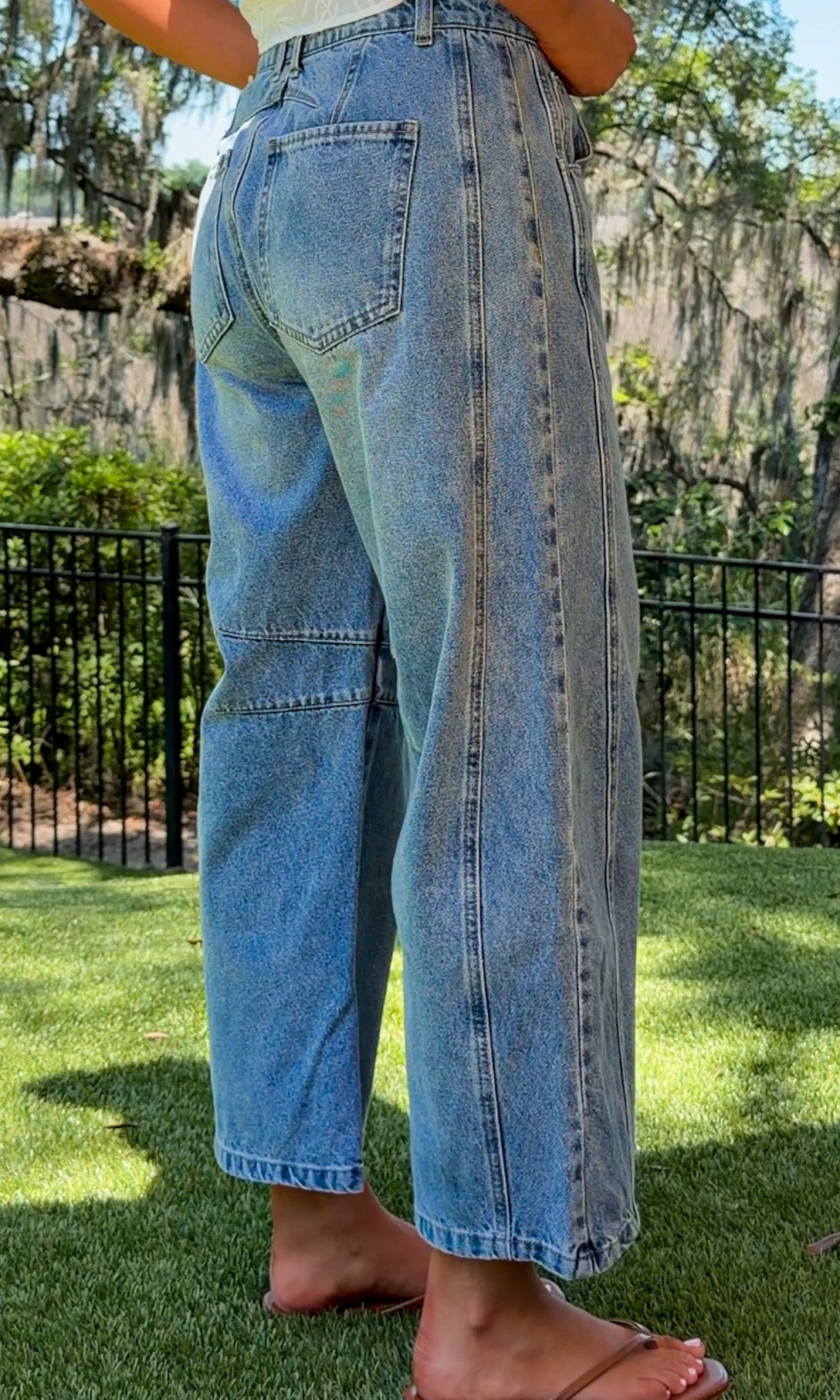 Person wearing light blue jeans standing on grass with trees and a fence in the background