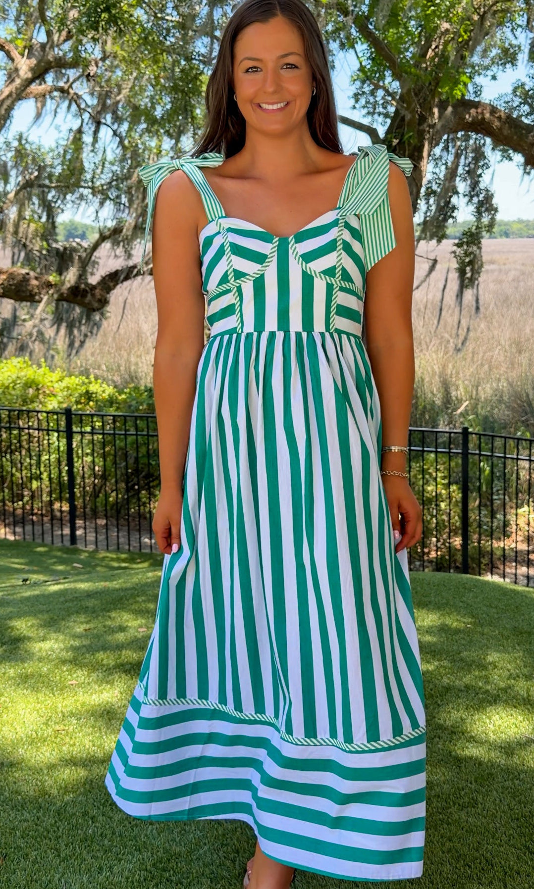Woman wearing a green and white striped dress standing outdoors with trees and grass in the background