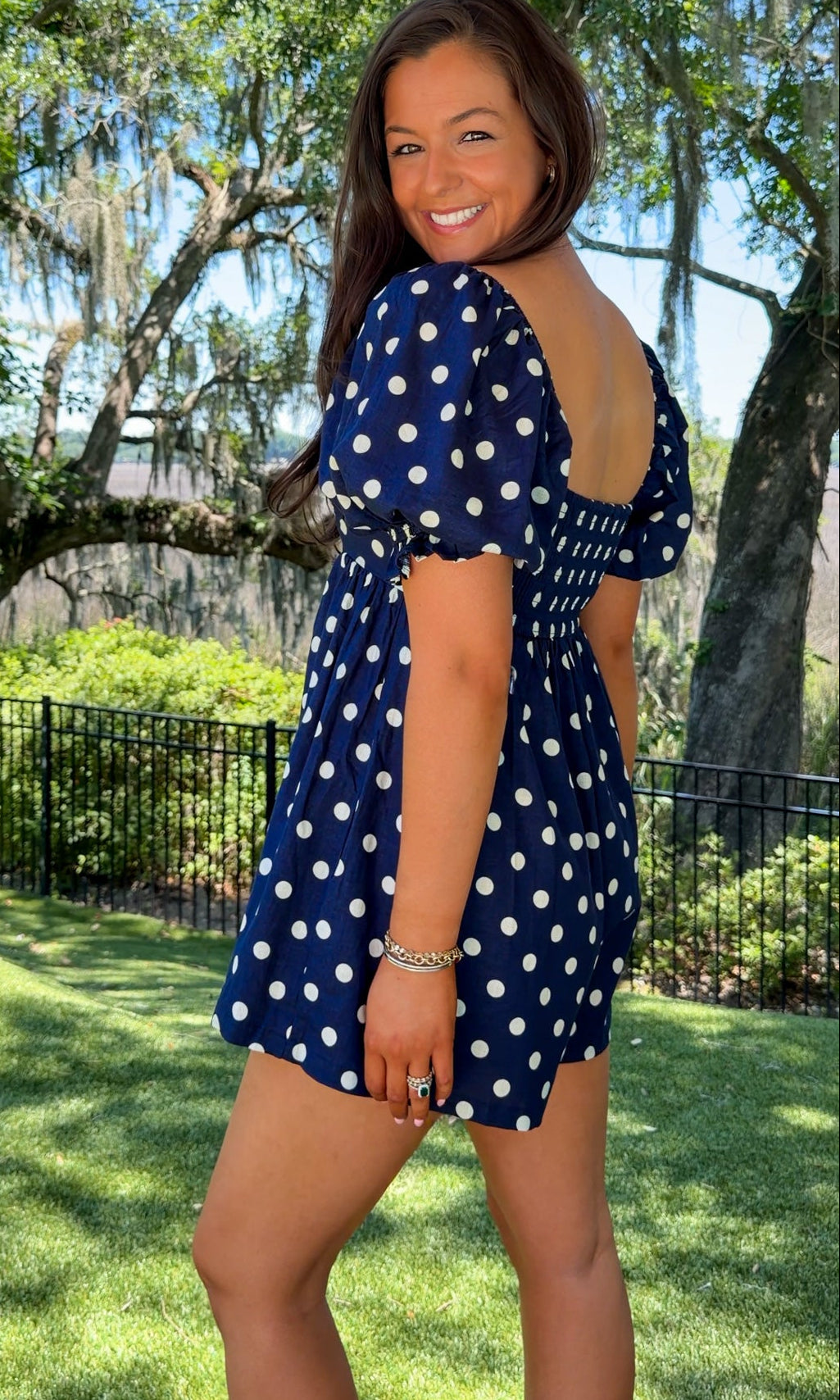 Woman in a blue polka dot dress standing outdoors with trees and grass in the background