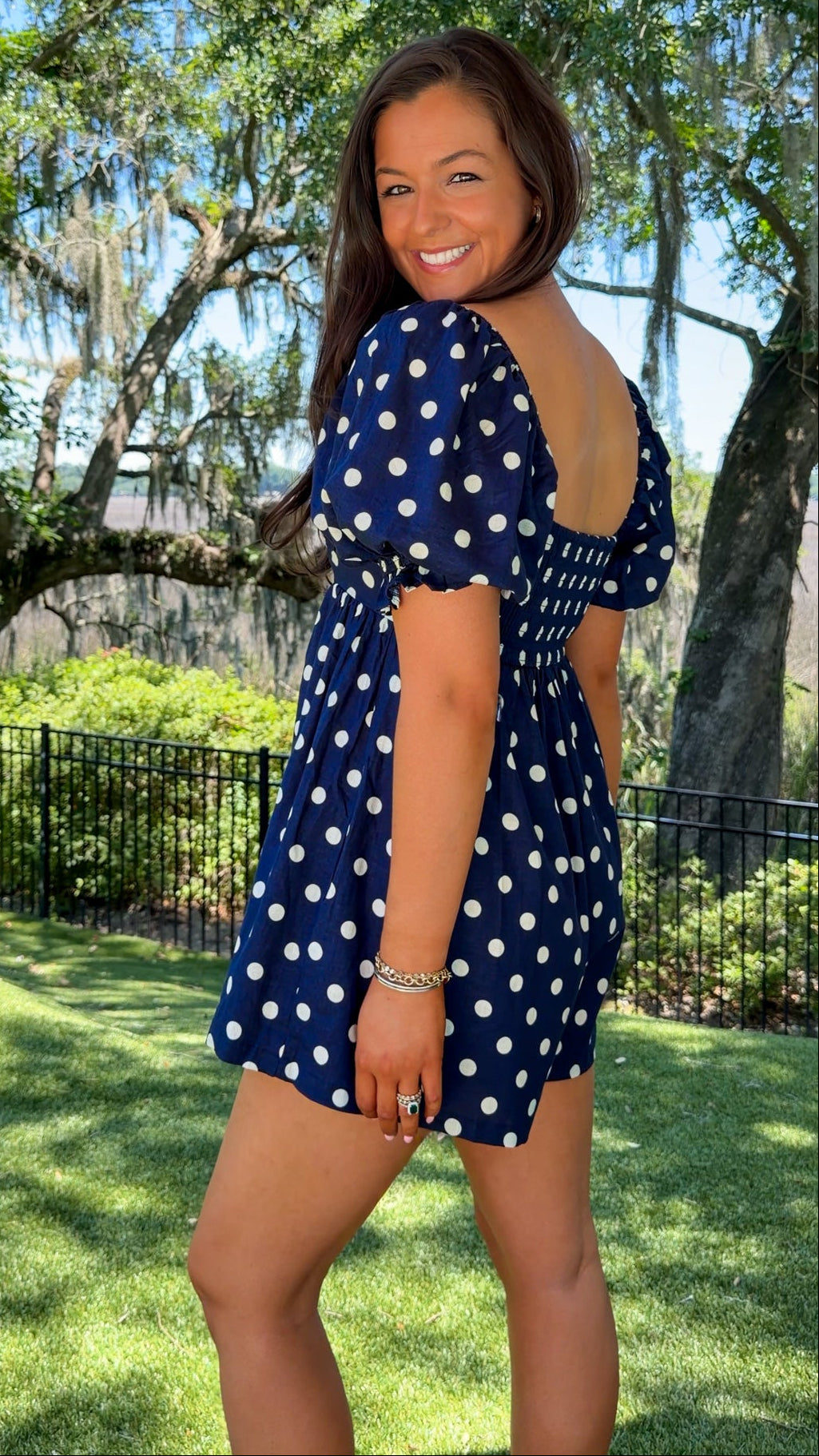 Woman in a blue polka dot dress standing outdoors with trees and grass in the background