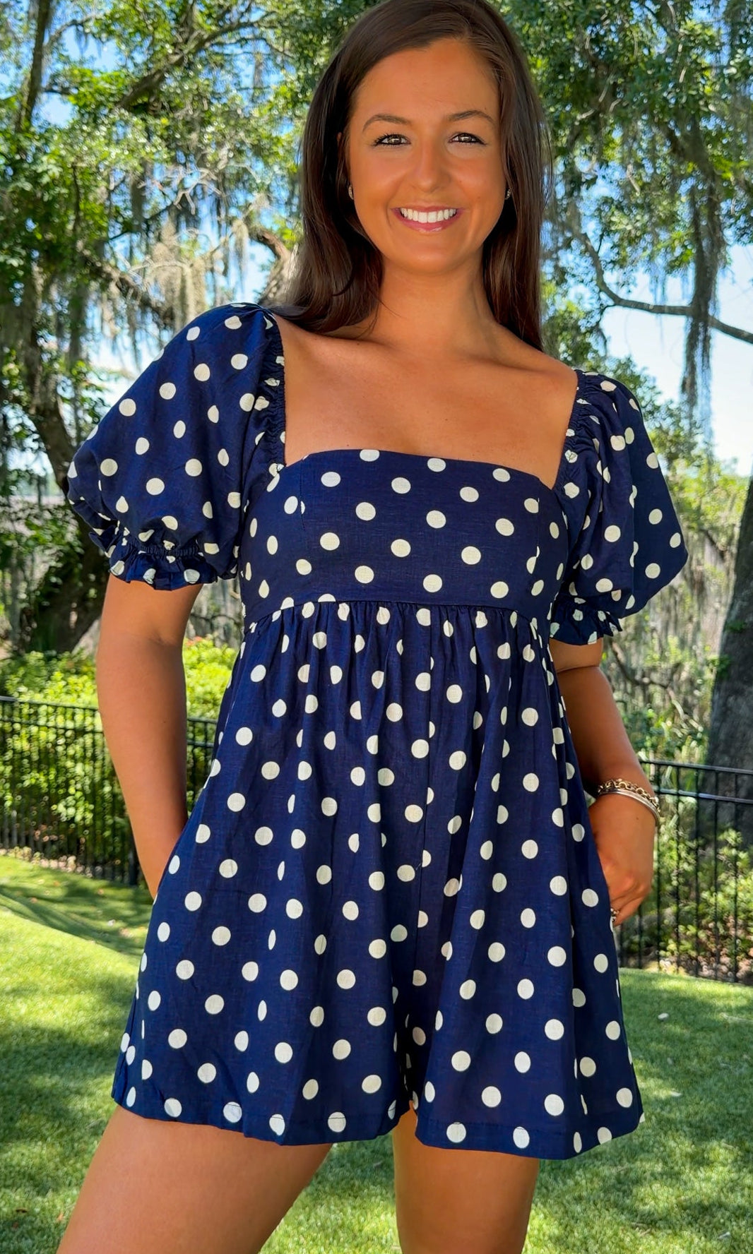 Woman wearing a blue polka dot dress standing outdoors with trees in the background