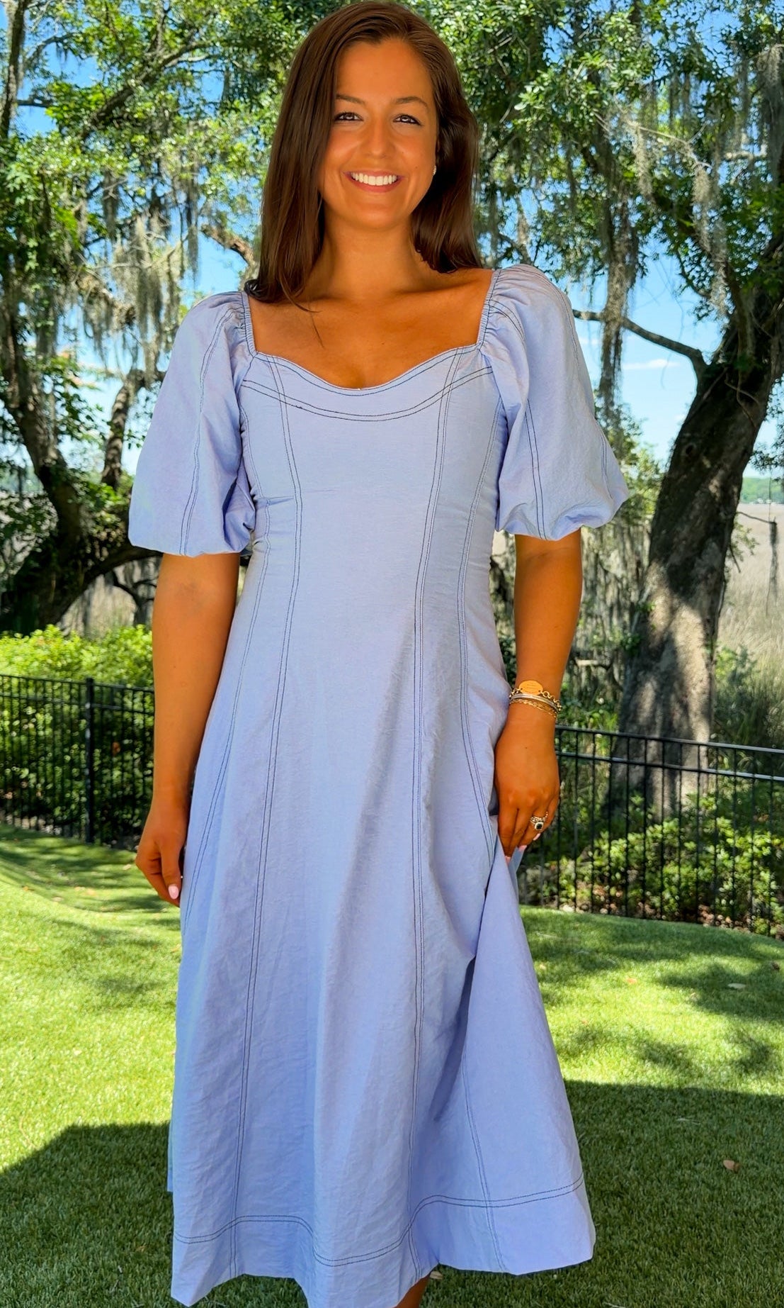 Woman in a light blue dress standing outdoors with trees and grass in the background