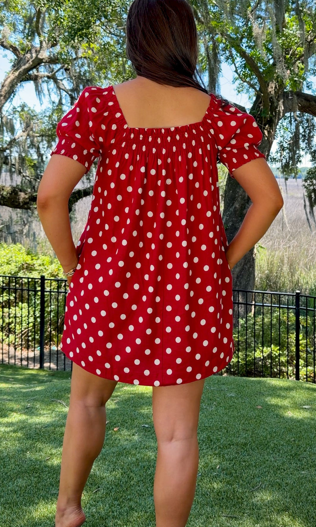 Person wearing a red polka dot dress standing outdoors with trees and grass in the background