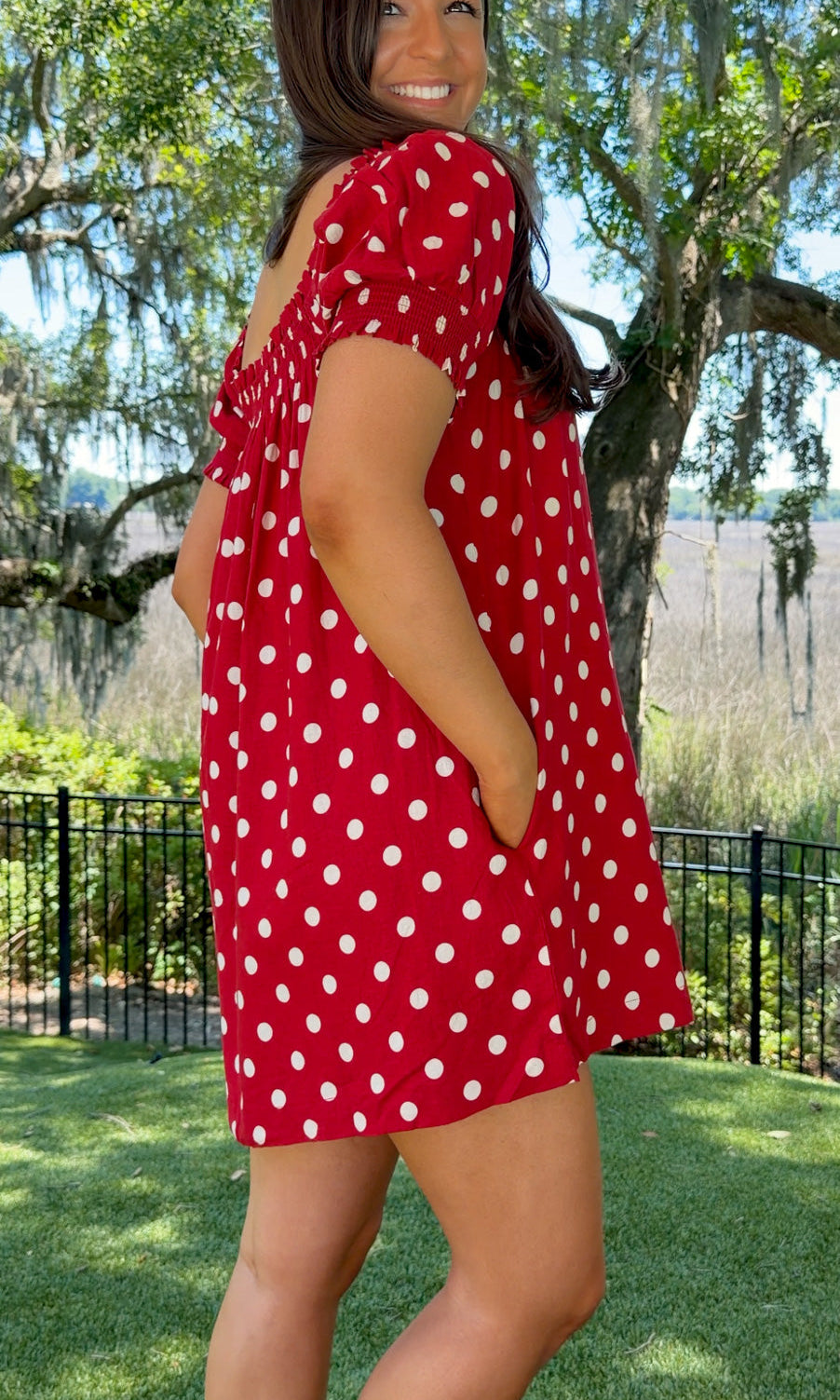 Woman wearing a red polka dot dress standing outdoors with trees and grass in the background