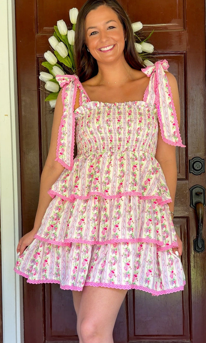 Pastel pink floral tiered mini dress with a smocked bodice, adjustable shoulder tie straps, lace trim detailing, and a flowy tiered skirt.