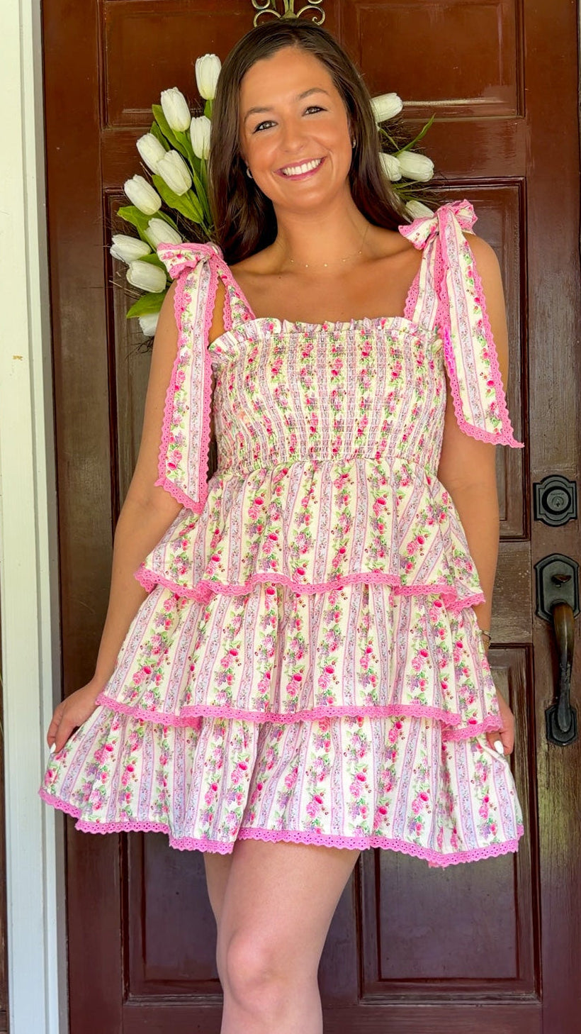 Pastel pink floral tiered mini dress with a smocked bodice, adjustable shoulder tie straps, lace trim detailing, and a flowy tiered skirt.