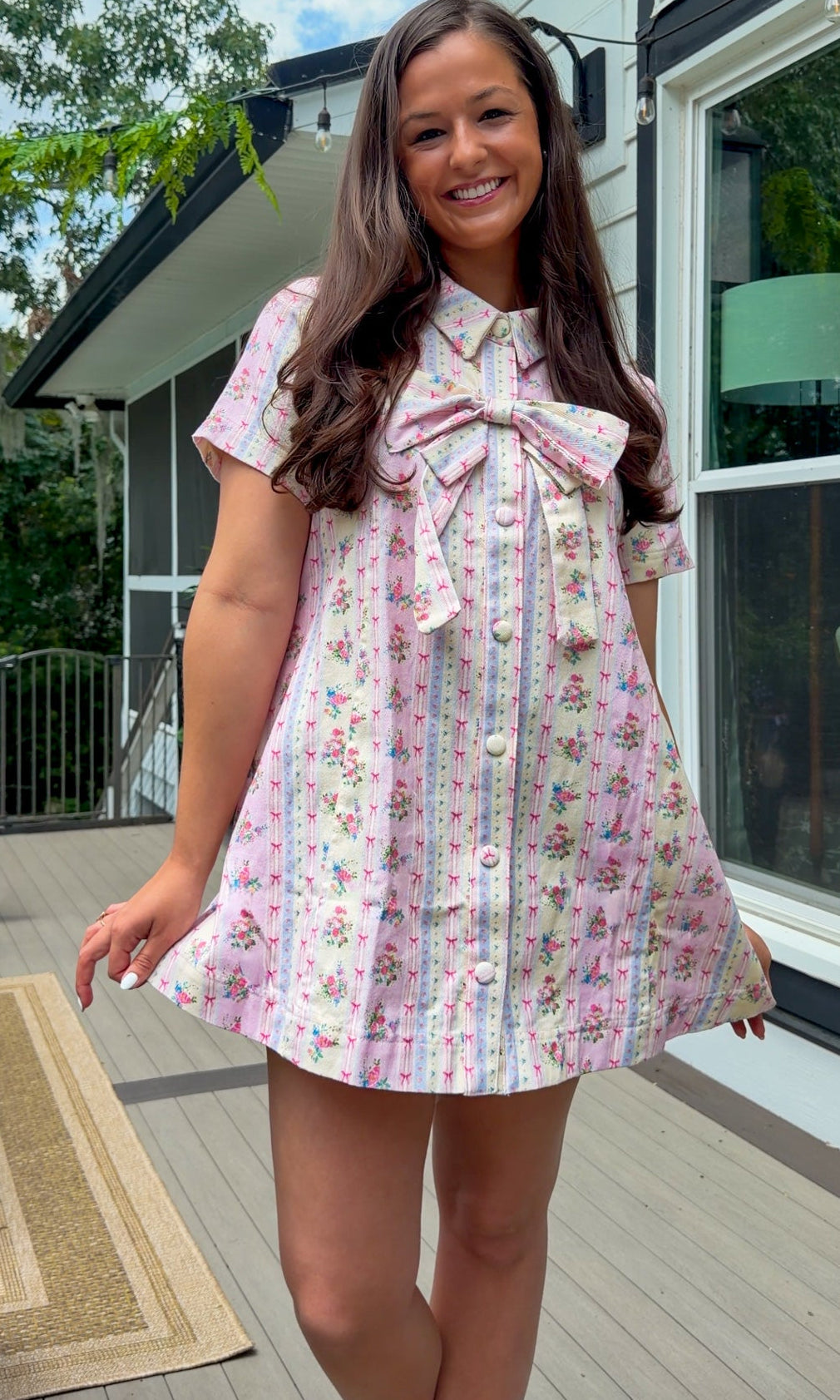 Light pink floral stripe mini dress with a collared neckline, front button closure, short sleeves, oversized bow detail, and relaxed A-line silhouette.