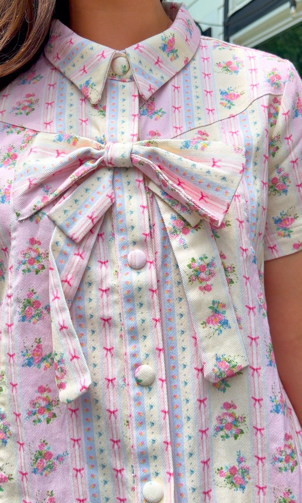 Light pink floral stripe mini dress with a collared neckline, front button closure, short sleeves, oversized bow detail, and relaxed A-line silhouette.