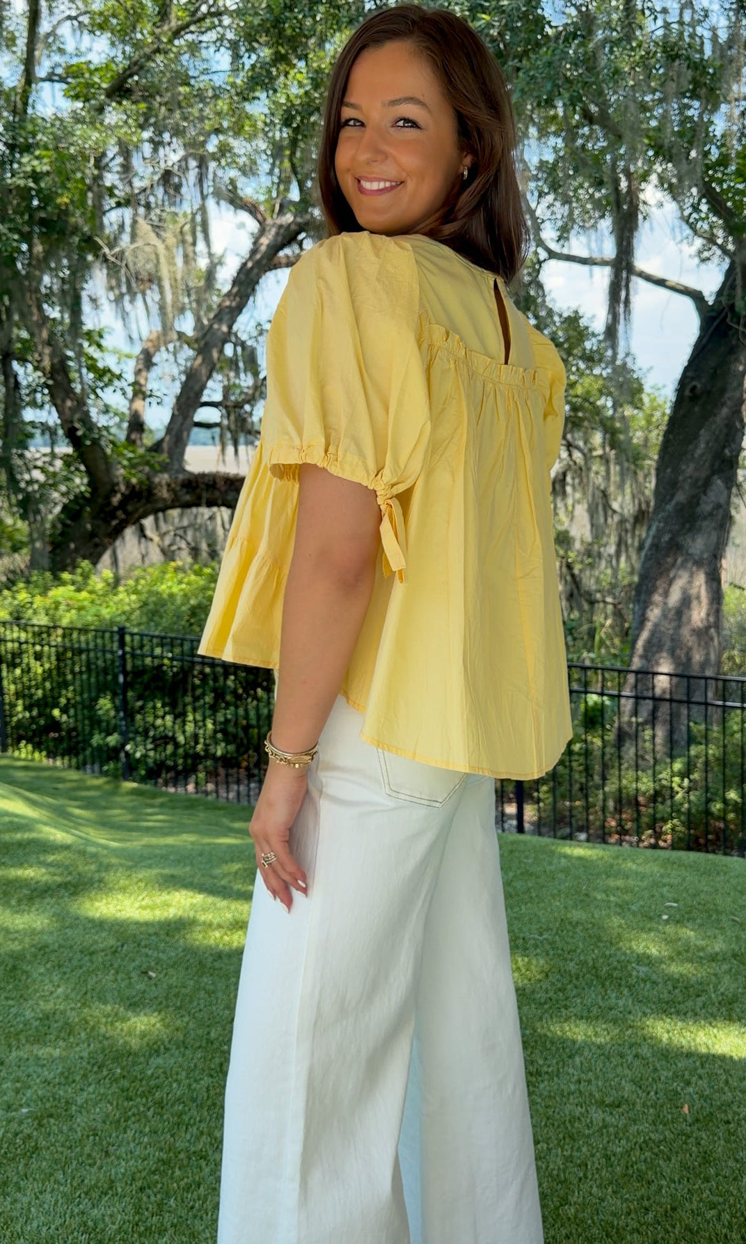 Butter yellow babydoll top featuring a round neckline, tiered shirring, short puff sleeves with adjustable tie cuffs, a ruffle-trimmed back, keyhole button closure, and a relaxed semi-cropped silhouette.