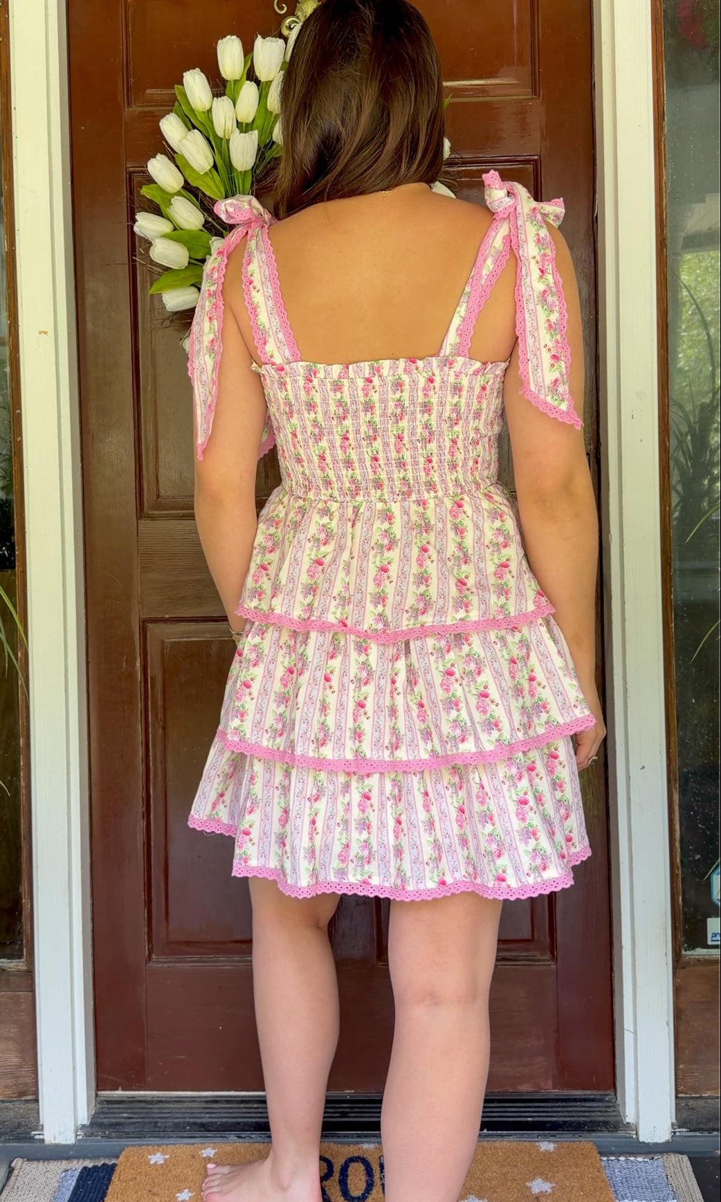 Pastel pink floral tiered mini dress with a smocked bodice, adjustable shoulder tie straps, lace trim detailing, and a flowy tiered skirt.