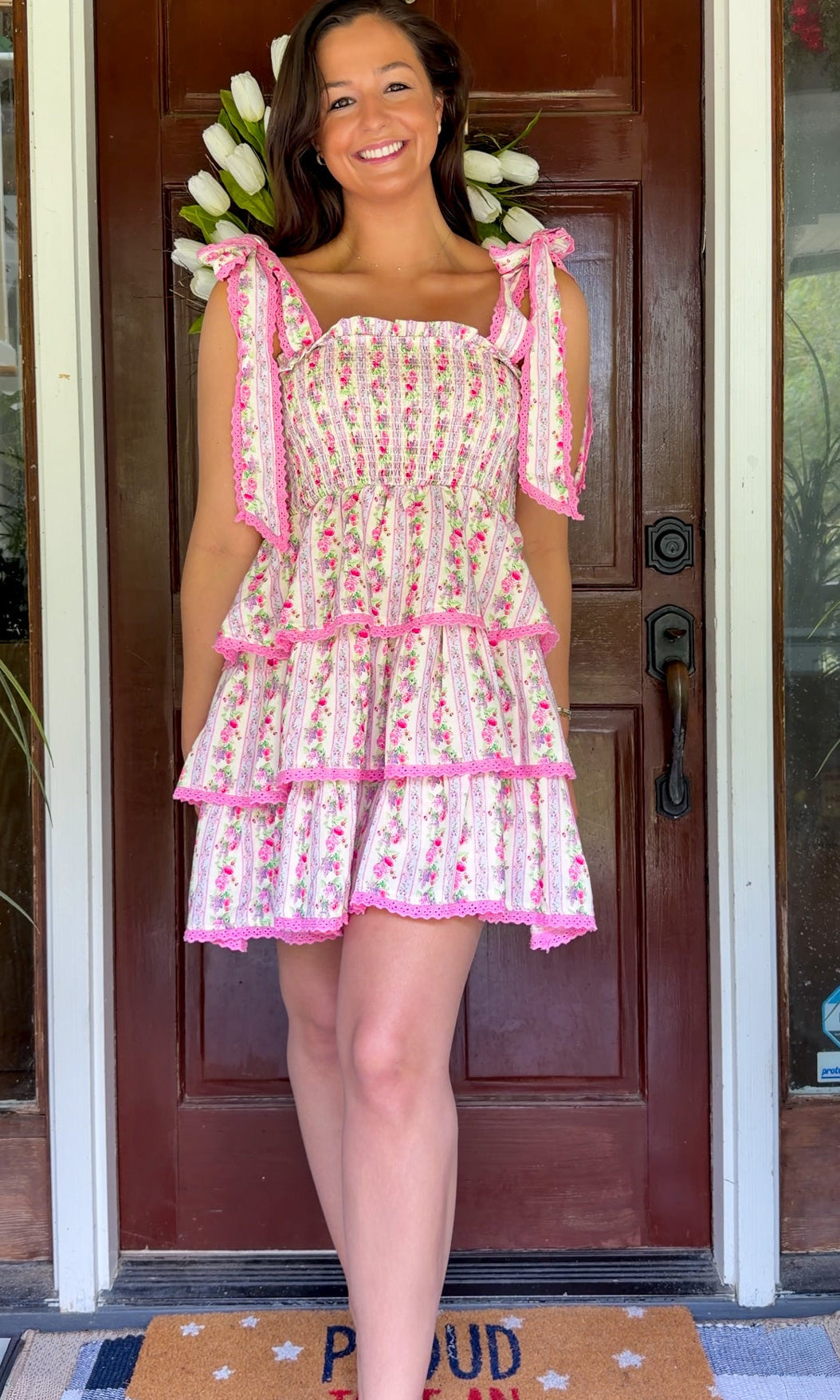 Pastel pink floral tiered mini dress with a smocked bodice, adjustable shoulder tie straps, lace trim detailing, and a flowy tiered skirt.