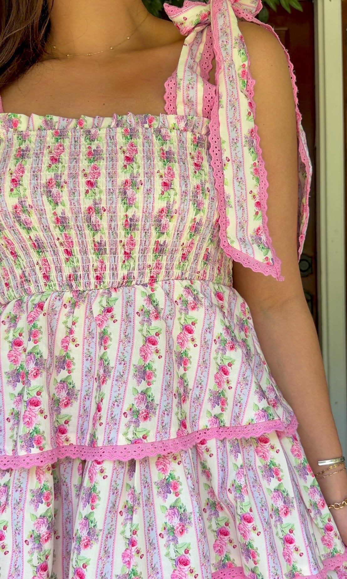 Pastel pink floral tiered mini dress with a smocked bodice, adjustable shoulder tie straps, lace trim detailing, and a flowy tiered skirt.
