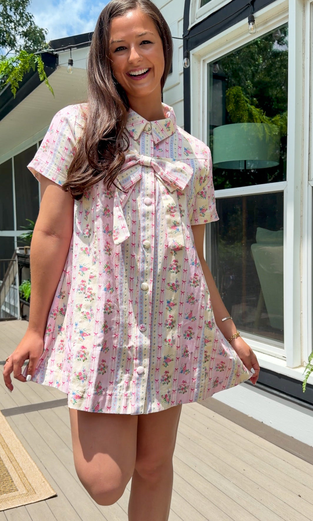 Light pink floral stripe mini dress with a collared neckline, front button closure, short sleeves, oversized bow detail, and relaxed A-line silhouette.