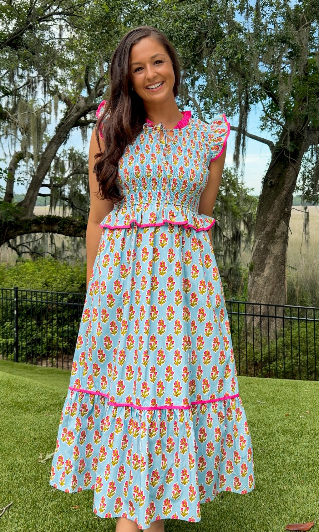 Light blue floral print midi dress featuring a fully smocked bodice, flutter sleeves with pink contrast trim, a keyhole tie neckline, ruffled peplum waist, tiered midi skirt, and side pockets.