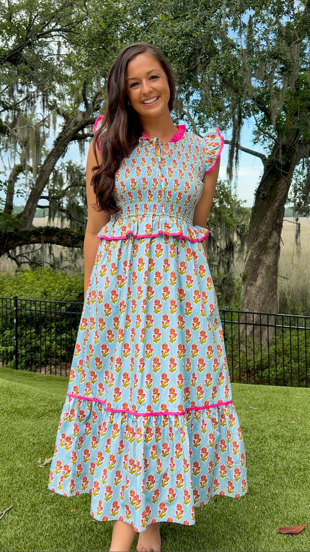 Light blue floral print midi dress featuring a fully smocked bodice, flutter sleeves with pink contrast trim, a keyhole tie neckline, ruffled peplum waist, tiered midi skirt, and side pockets.